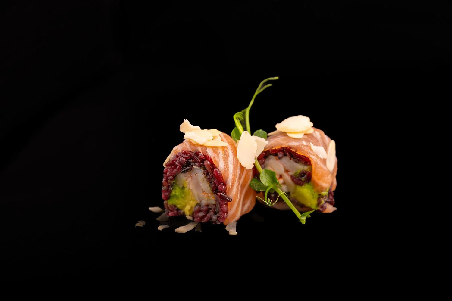 Zwei Sushi-Rollen mit Lachs, Avocado, rote Bohnen und Garnelen auf schwarzem Hintergrund, garniert mit Kresse und geriebenem Käse.