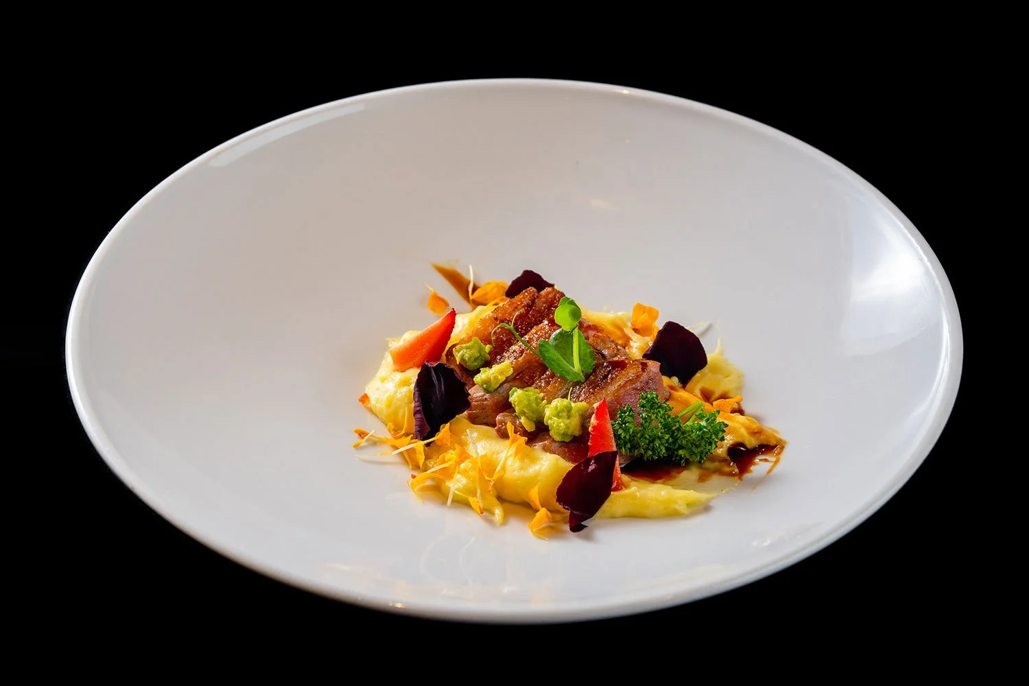 Feines, künstlerisch angerichtetes Gericht mit Kartoffelpüree, Speck, Gemüse und Kräutern auf weißem Teller vor schwarzem Hintergrund.