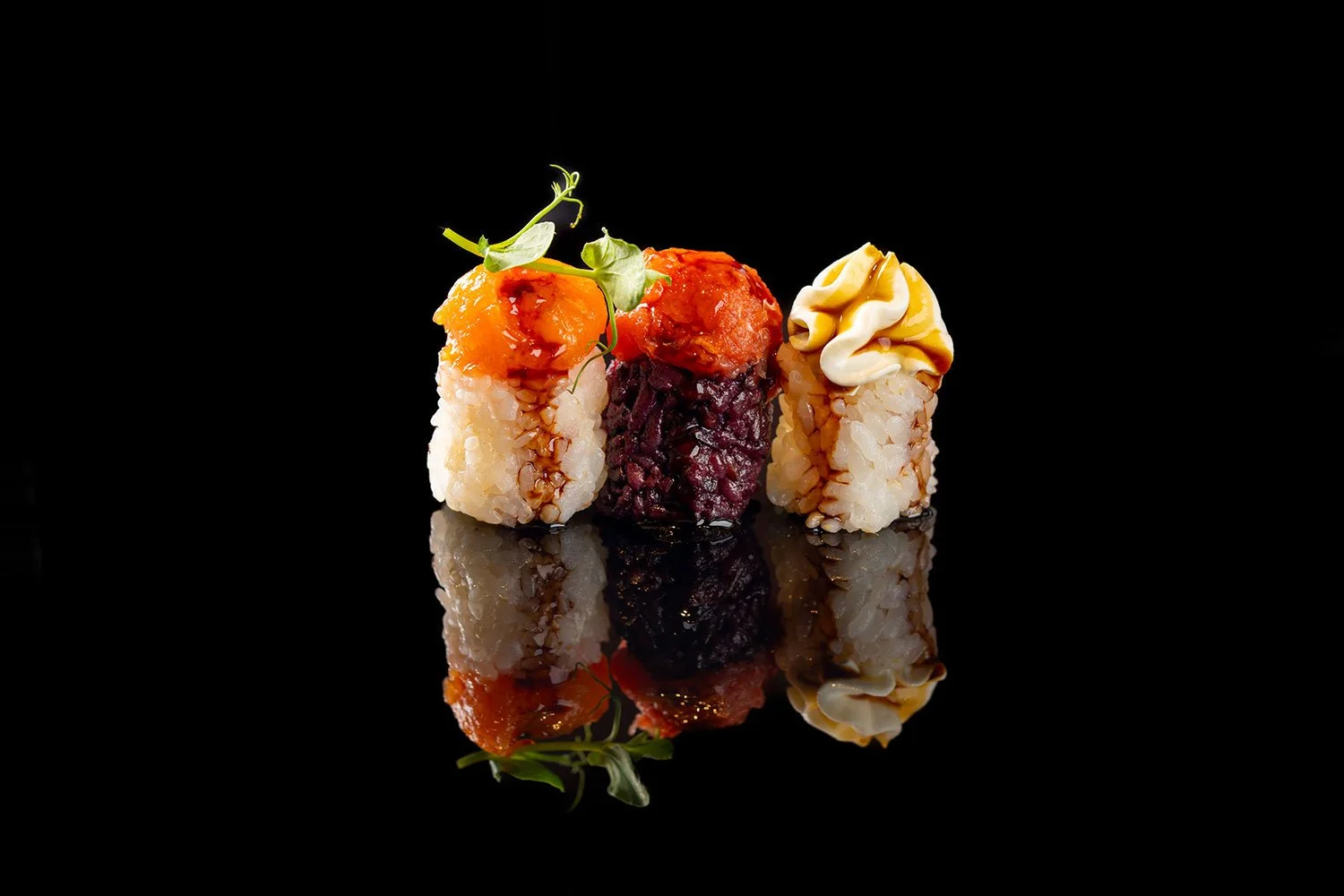 Drei Sushi-Rollen auf schwarz-reflektierender Oberfläche, mit verschiedenen Toppings und Garnieren.