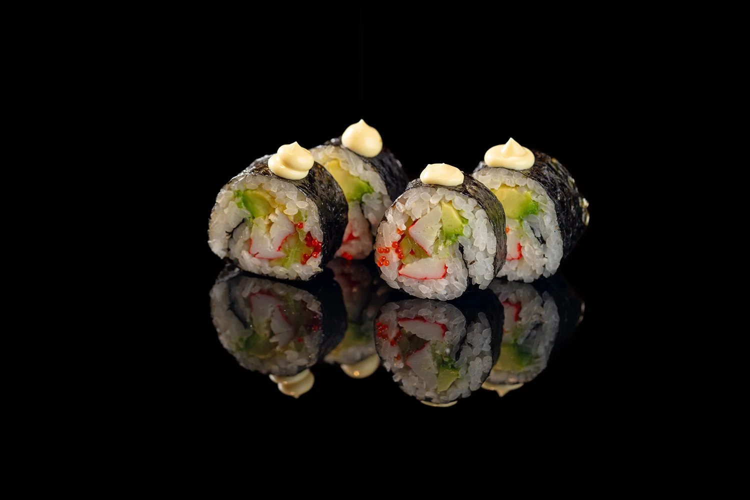 Vier Sushi-Rollen mit Avocado, Krabben und Mayonnaise auf glatter schwarzer Oberfläche, mit deren Spiegelbild im Vordergrund.
