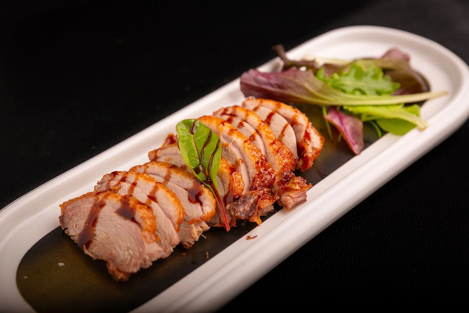 In diesem Bild ist ein Teller mit in Scheiben geschnittenem, gebratenem Fleisch, garniert mit einer grünen Blattform, und serviert mit gemischtem Salat auf einer weißen, länglichen Schale.