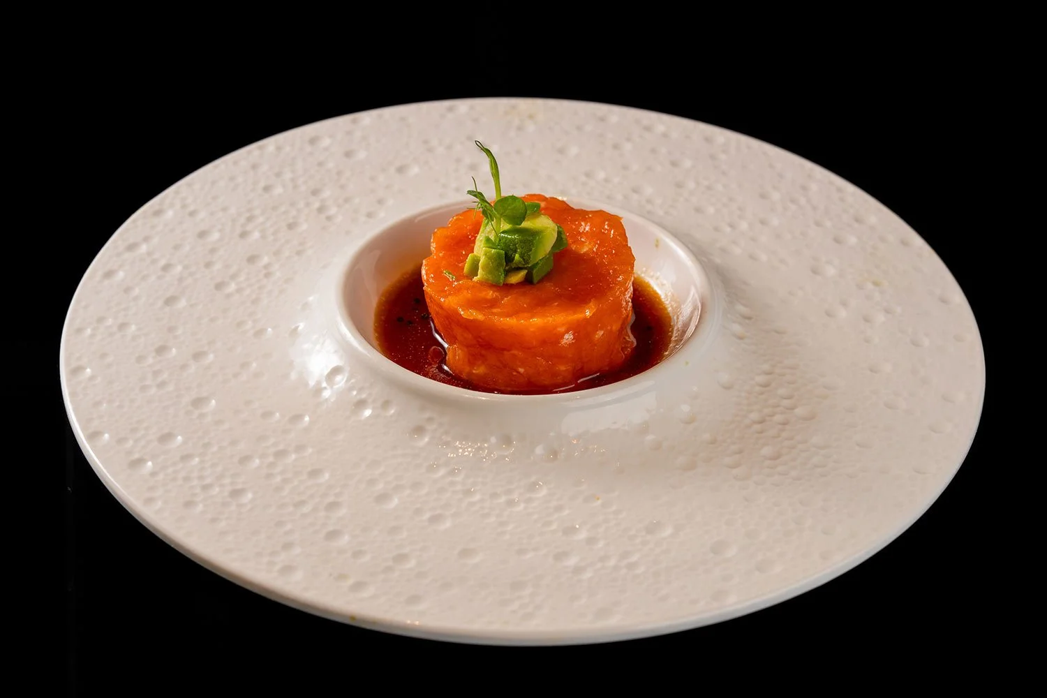 Ein Teller mit einer kleinen Portion saftiger, orangefarbener Fisch-Tatar, garniert mit kleinen Avocadostücken und einem grünen Kräuterblatt, serviert in einem weißen, einzigartigen Speiseteller