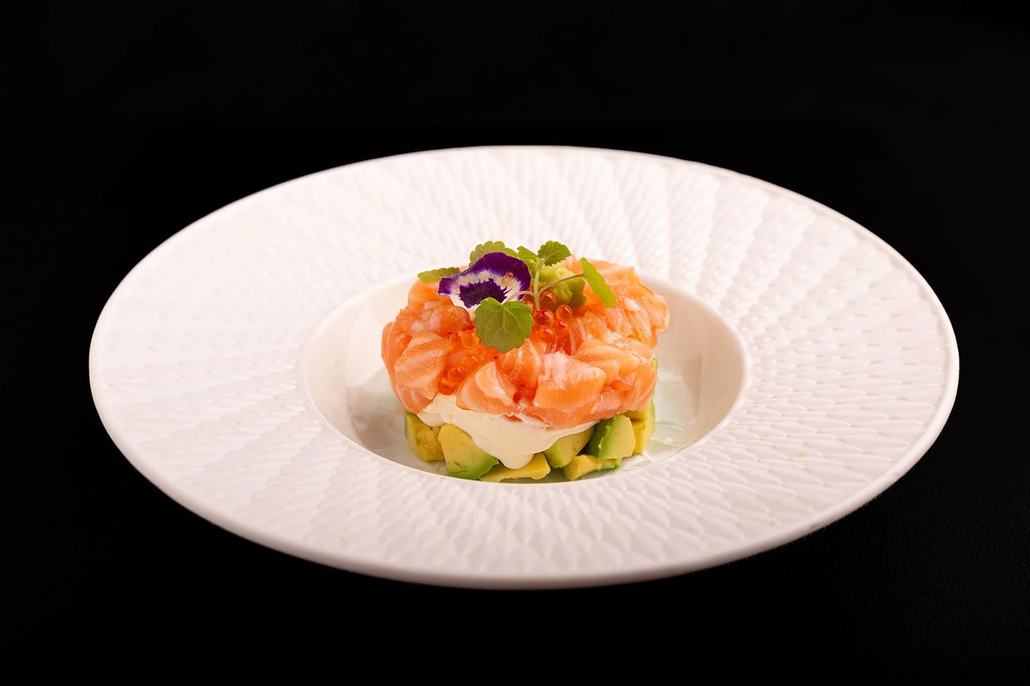 Eleganter Teller mit Lachs-Tartar, Avocado, Ei und Garnele, garniert mit Blättern und essbaren Blumen, auf einer weißen Platte