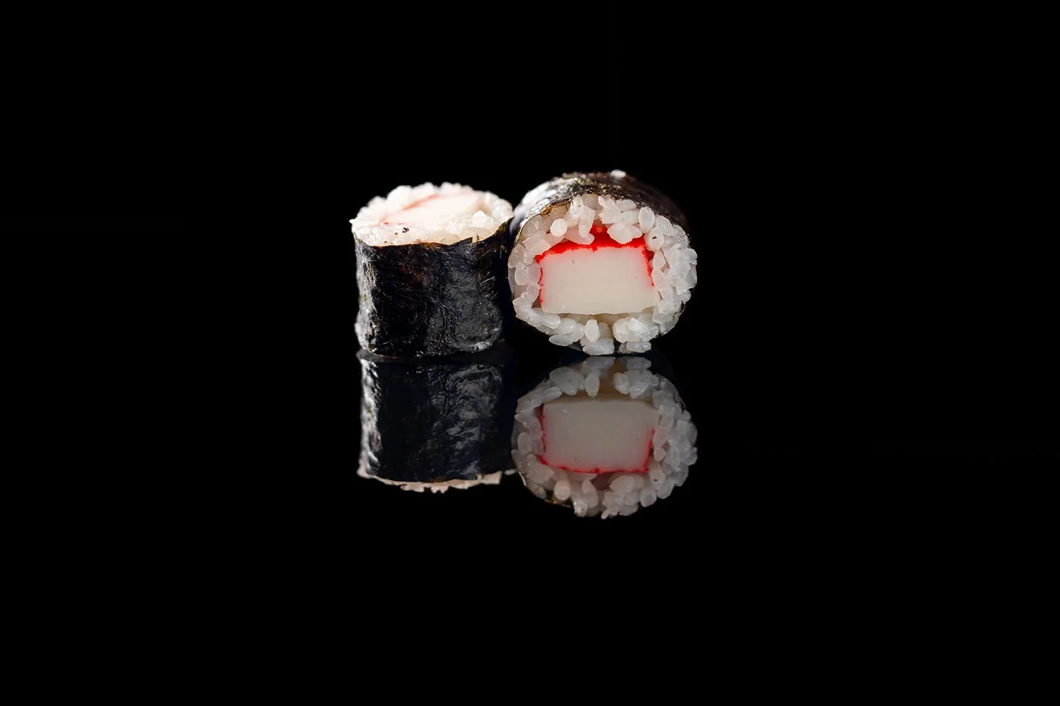 Zwei Sushi-Rollen mit Nori, Reis und Krabbenfleisch vor schwarzem Hintergrund, die eine Reflexion auf einer spiegelnden Oberfläche zeigen.