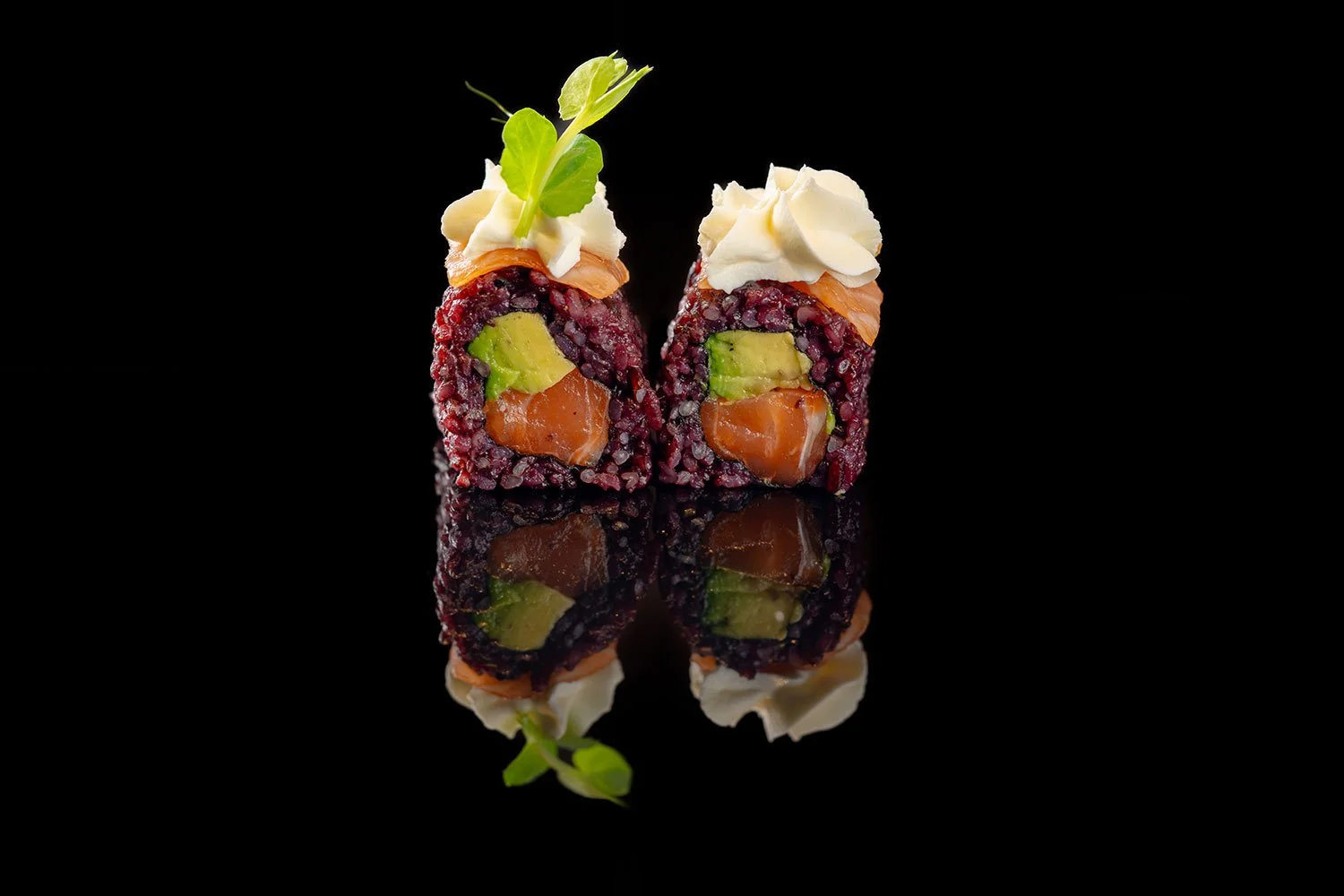 Zwei Sushi-Rollen mit Lachs, Avocado und schwarzem Reis, garniert mit Sahne und kleinen grünen Kräutern, vor schwarzem Hintergrund mit Spiegelung.