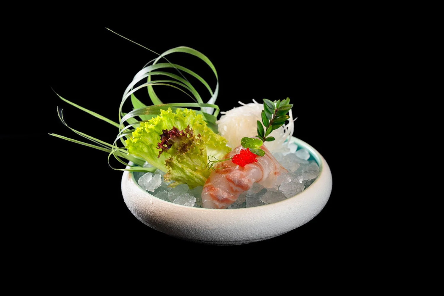 Sashimi mit Garnelen, Salat, Limone, Roteise, und Kräutern auf Eis in einer weißen Schüssel gegen schwarzen Hintergrund.