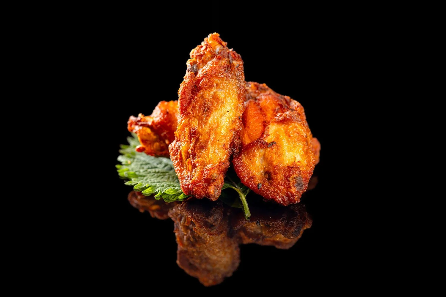 Zwei frittierte Chicken Wings auf einem grünen Blatt vor einem schwarzen Hintergrund, mit Reflexion auf der Oberfläche.