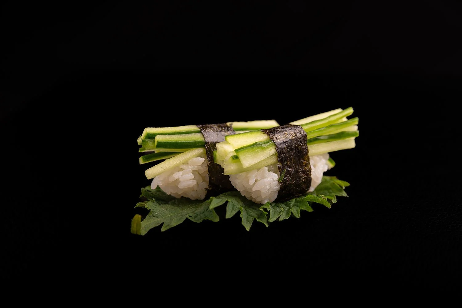 Sushi mit Gurken, Noriblatt, Reis und einem grünen Blatt auf schwarzem Hintergrund.