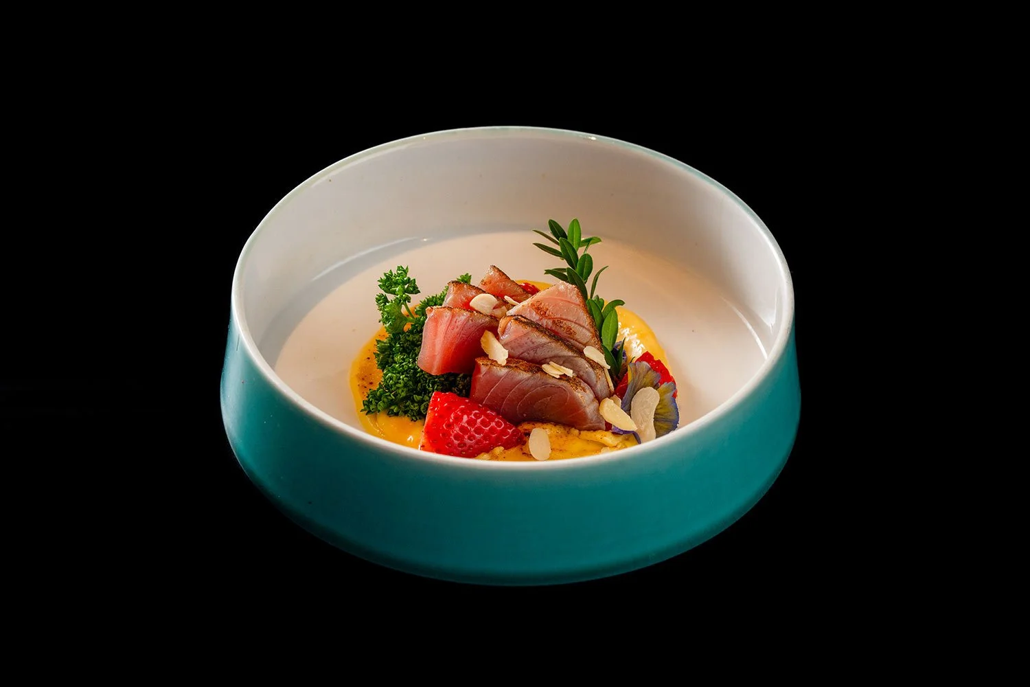 Feines Gericht mit rohem Fisch, Beeren, Kräutern und Mandelblättchen, serviert in einer blauen Schüssel auf schwarzem Hintergrund.