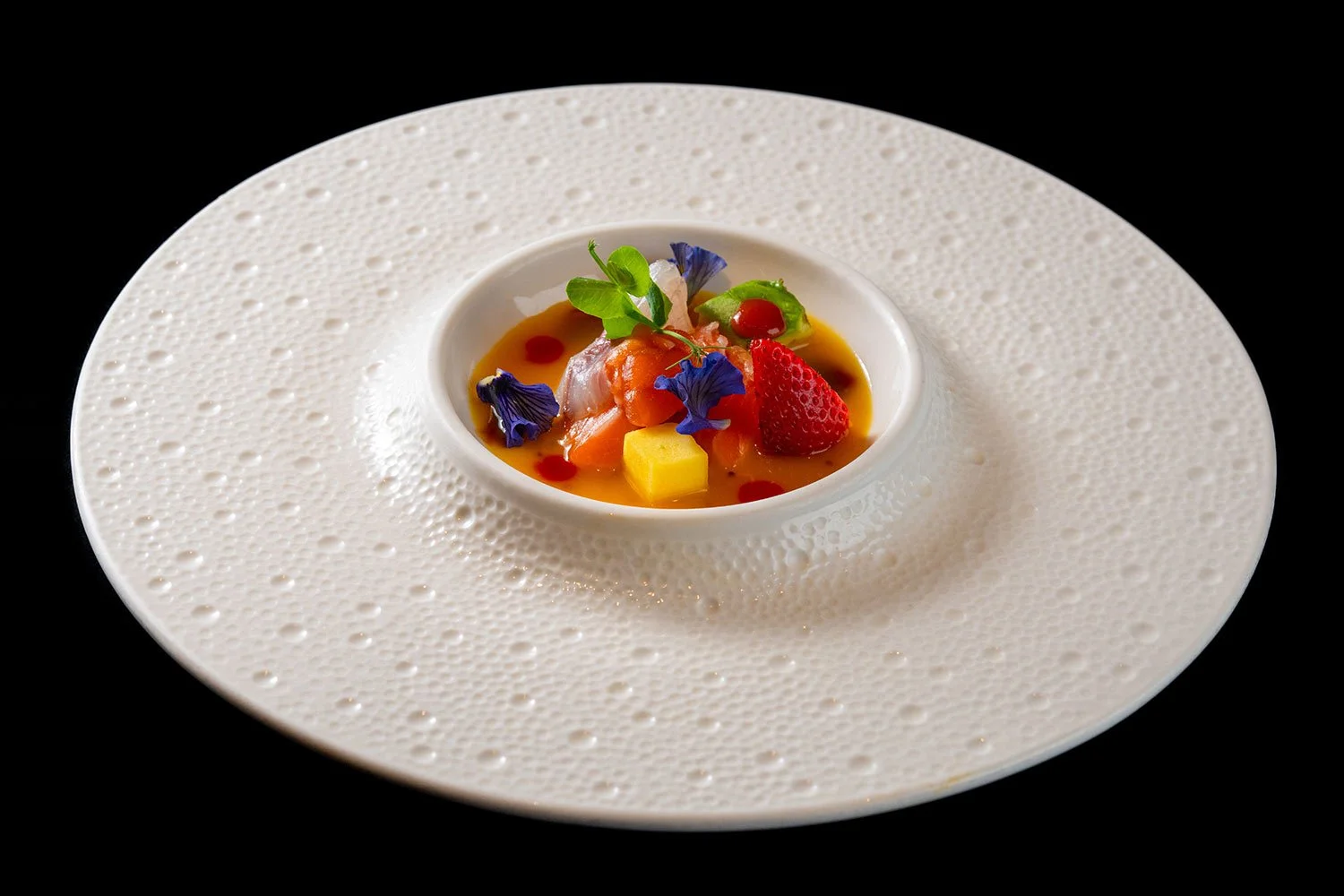 Elegantes Dessert mit Beeren, Mango, Gurke, Blüten und Sauce in einer weißen Schale auf einem großen weißen Teller mit strukturierter Oberfläche auf schwarzem Hintergrund.