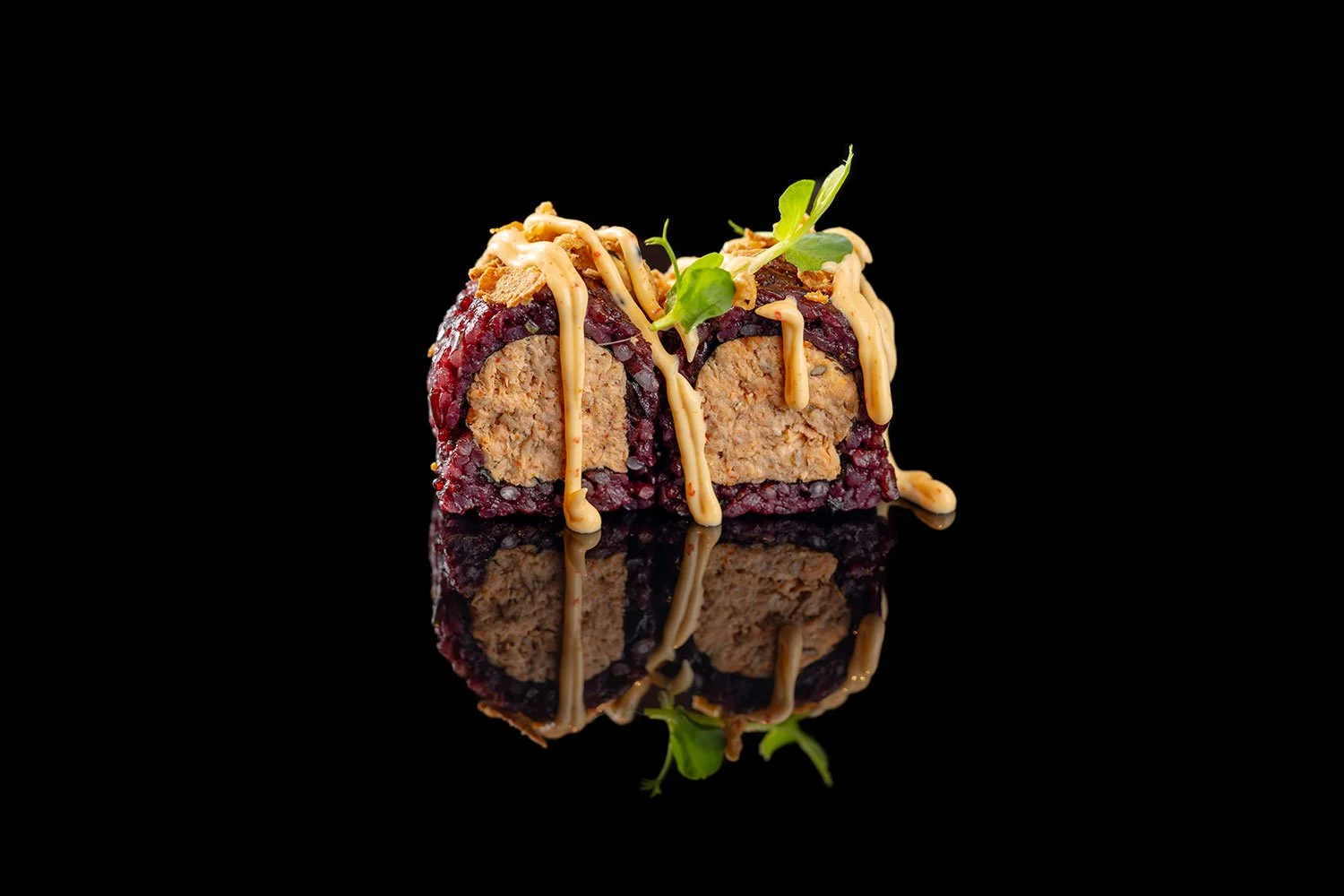 Ein Sushi-Roll mit Programm, darunter Gurken und krustige Krümel, garniert mit einer cremigen Soße und kleinen grünen Blättern auf schwarzem Hintergrund, mit Spiegelung darunter.