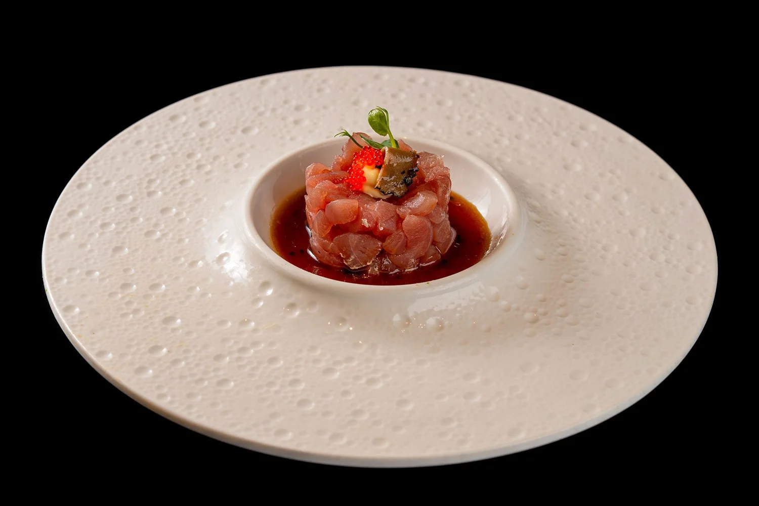 Rote Thunfisch-Tatar in einer kleinen weißen Schüssel, garniert mit einer grünen Kresseblüte, umgeben von einer roten Sauce auf einem großen, weißen, strukturierten Teller mit schwarzem Hintergrund.