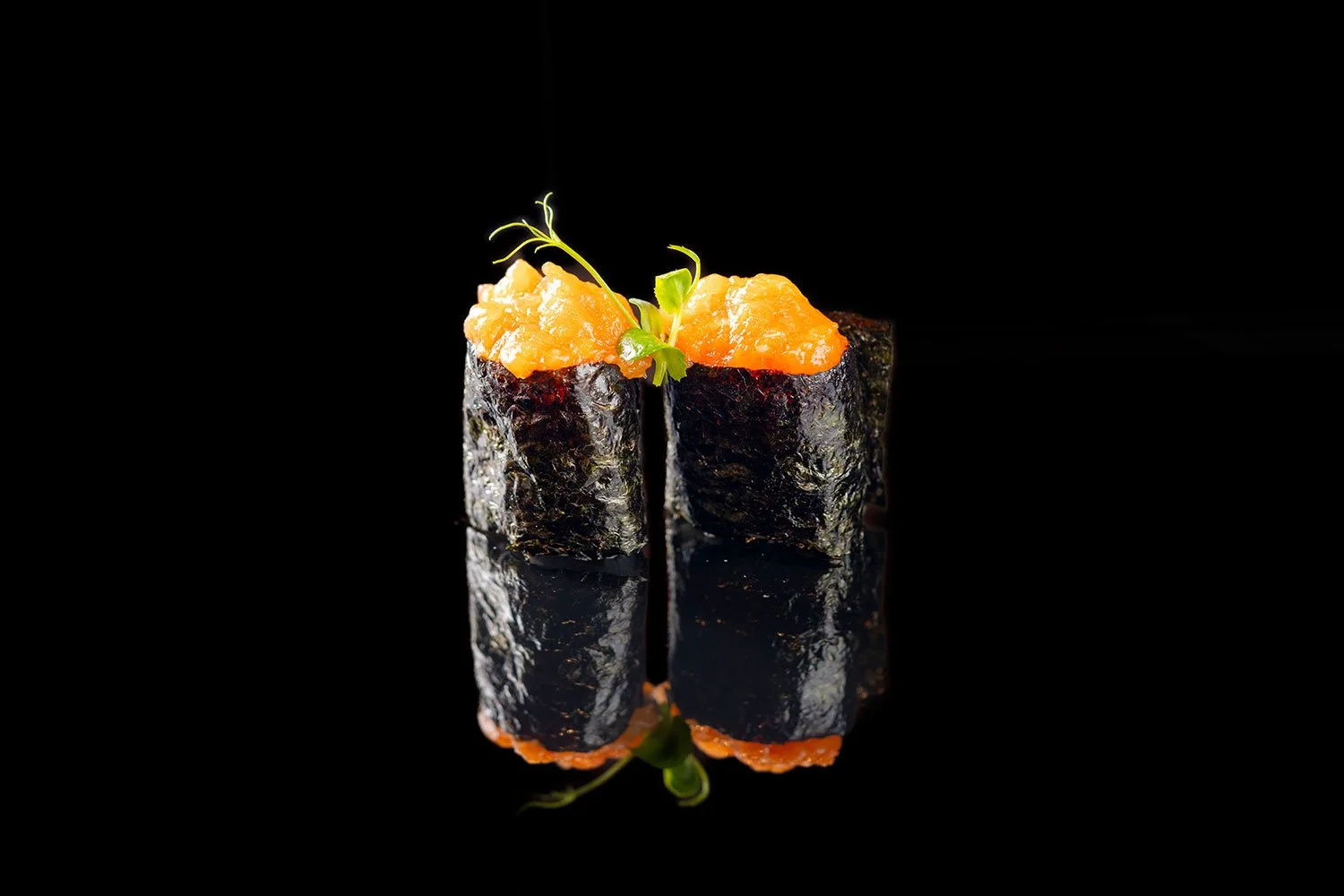 Zwei Sushi-Rollen mit schwarzer Nori-Alge, gefüllt mit Orangem, garniert mit grünen Sprossen, auf schwarzem Hintergrund mit Reflexion.