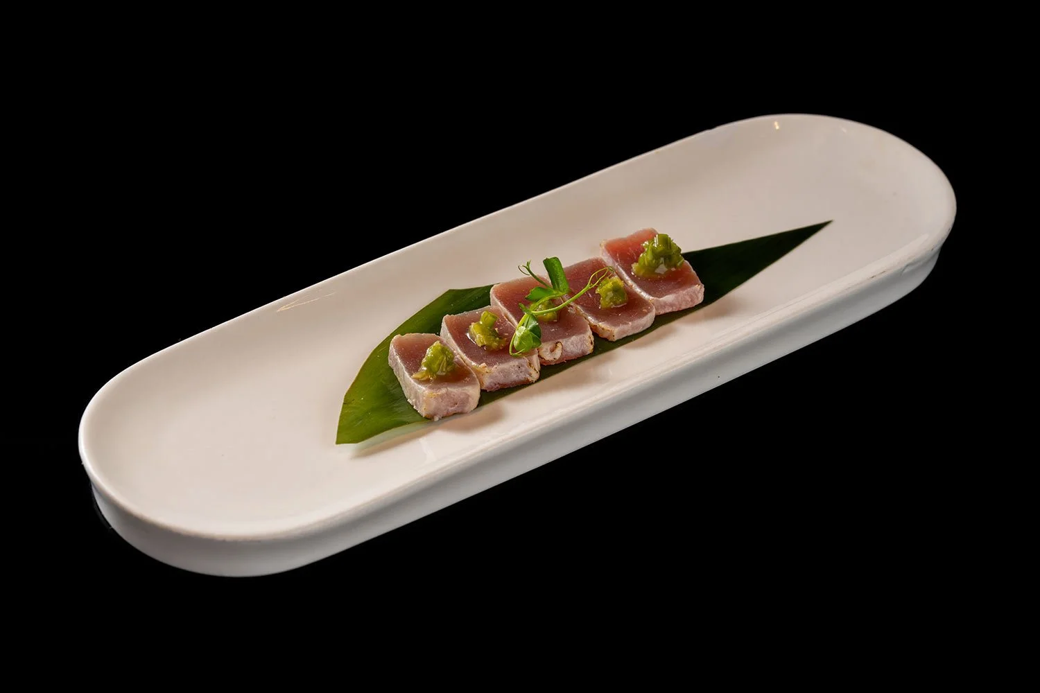 Auf einem weißen, ovalen Teller liegen fünf Scheiben rohen Thunfisch mit grünen Garnelen und Kräutern auf einer grünen Blattschicht, gegen den schwarzen Hintergrund.