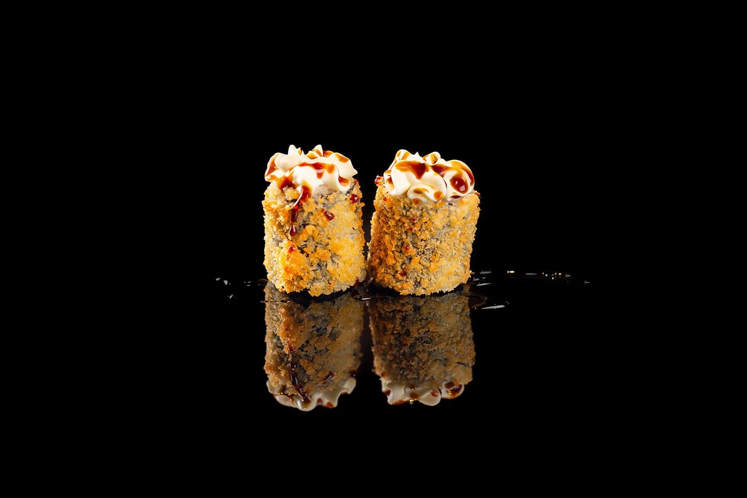 Zwei panierten Sushi-Rollen mit Frischkäse, Sauce und Mayonnaise auf einem schwarzen Hintergrund, mit reflektierendem Oberflächenspiegel.