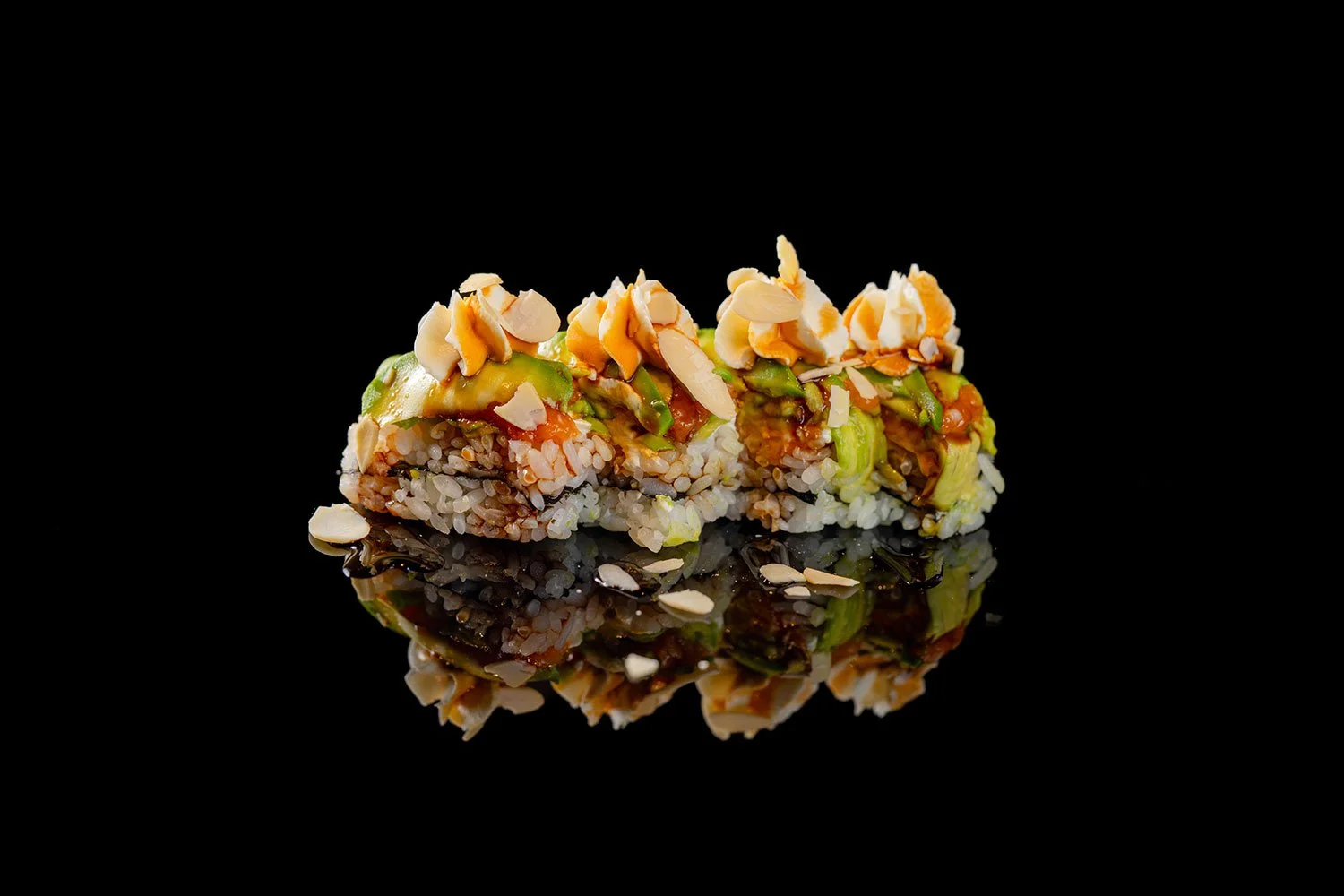 Nahaufnahme von Sushi-Rollen mit Avocado, Mandeln und Saucen auf schwarzem Hintergrund, Spiegelbild sichtbar