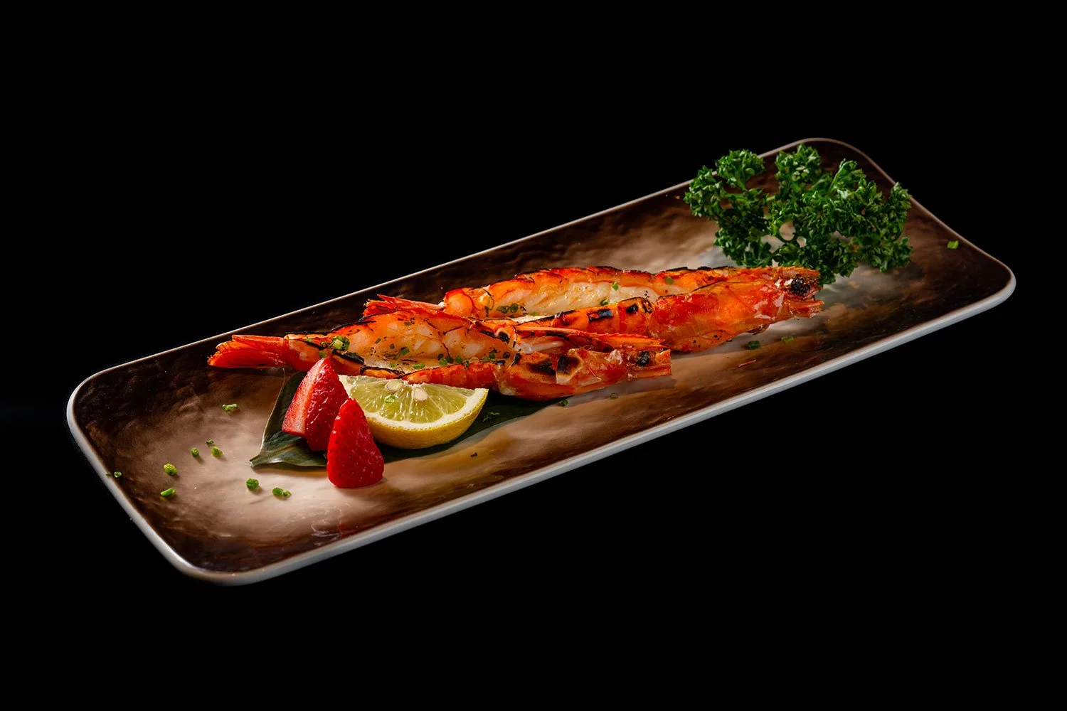 Gegrillte Garnelen auf einem schwarzen Teller mit Zitronenscheibe, Erdbeeren, süßer Chili-Sauce und Petersilie, schwarzer Hintergrund