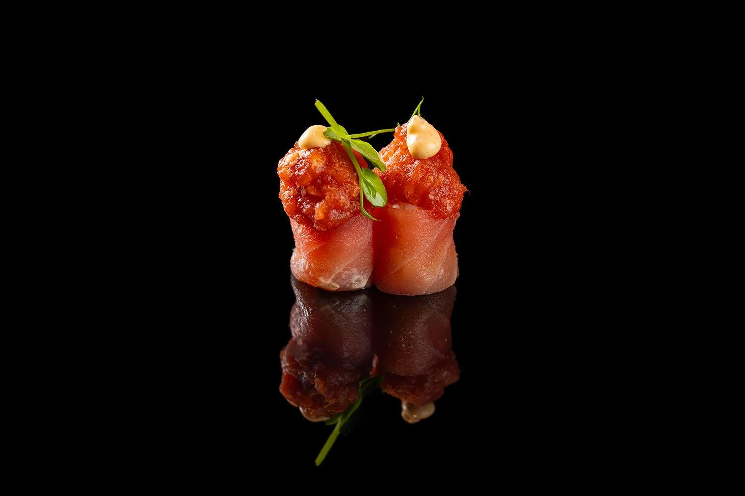 Zwei Sushi-Rollen mit roten Fischfleisch-Toppings, garniert mit grünen Blättern und cremiger Sauce, auf schwarzem Hintergrund mit Reflexion