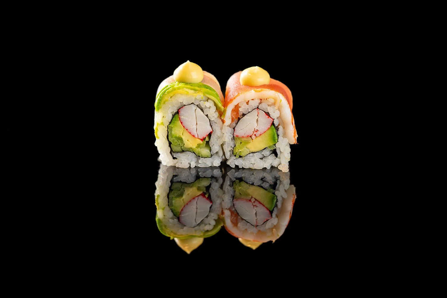 Zwei Sushi-Rollen mit Lachs, Avocado, Krabben und Gurke, eine mit grünem Fisch und die andere mit rotem Fisch, auf spiegelndem schwarzen Hintergrund