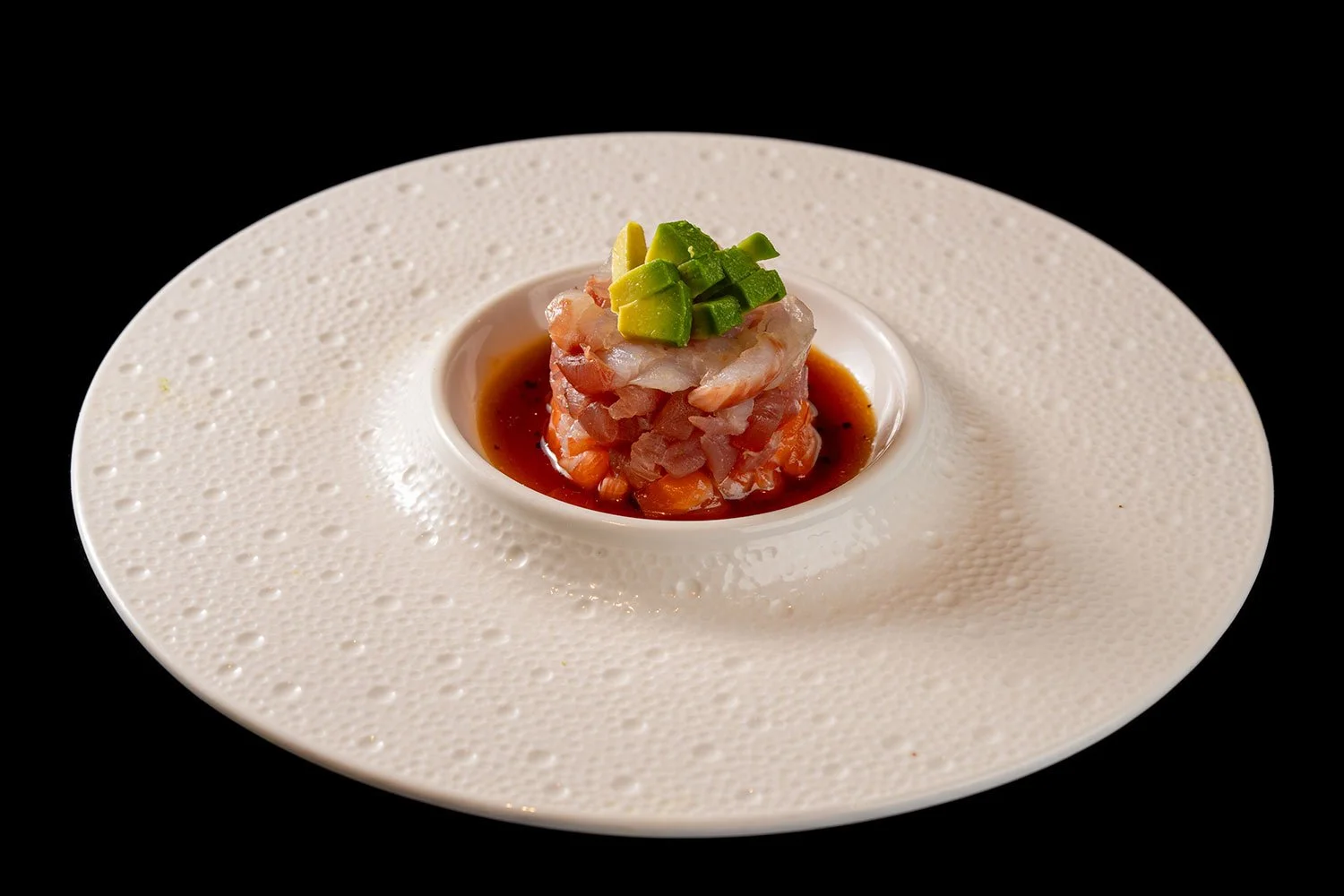 Vorspeise mit rohem Fisch, gewürfelte Avocado und Sojasauce auf einem weißen, vertieften, schwarzen Hintergrund