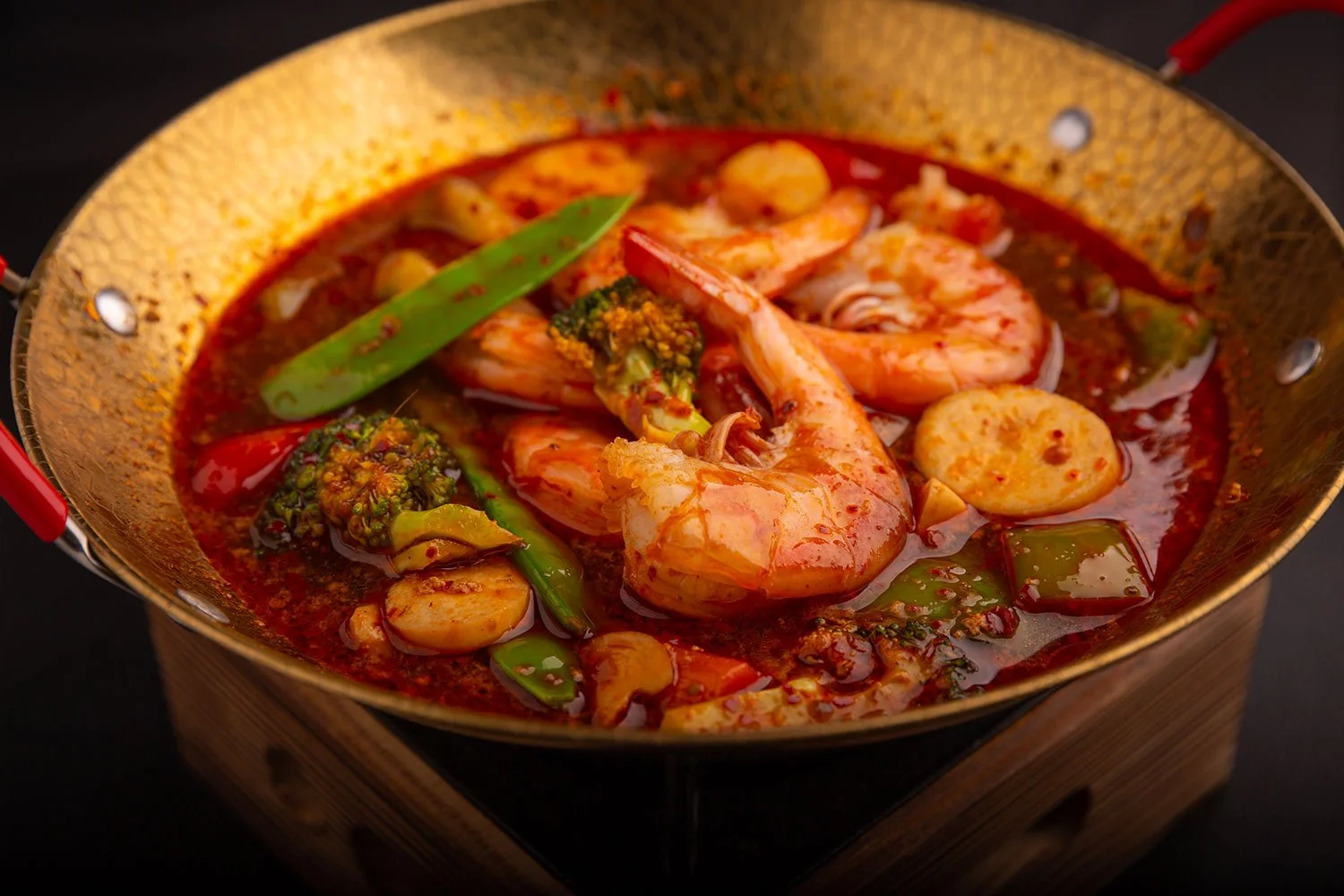 Ein Topf mit koreanischem Eintopf, gefüllt mit Garnelen, Brokkoli, grüner Paprika, Karotten, Zwiebeln und Chili in roter Brühe, auf dunklem Hintergrund.