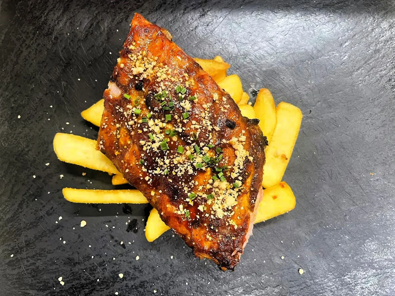 Gegrilltes Stück Fleisch auf Pommes Frites, garniert mit Kräutern und Parmesan auf einem schwarzen Teller.