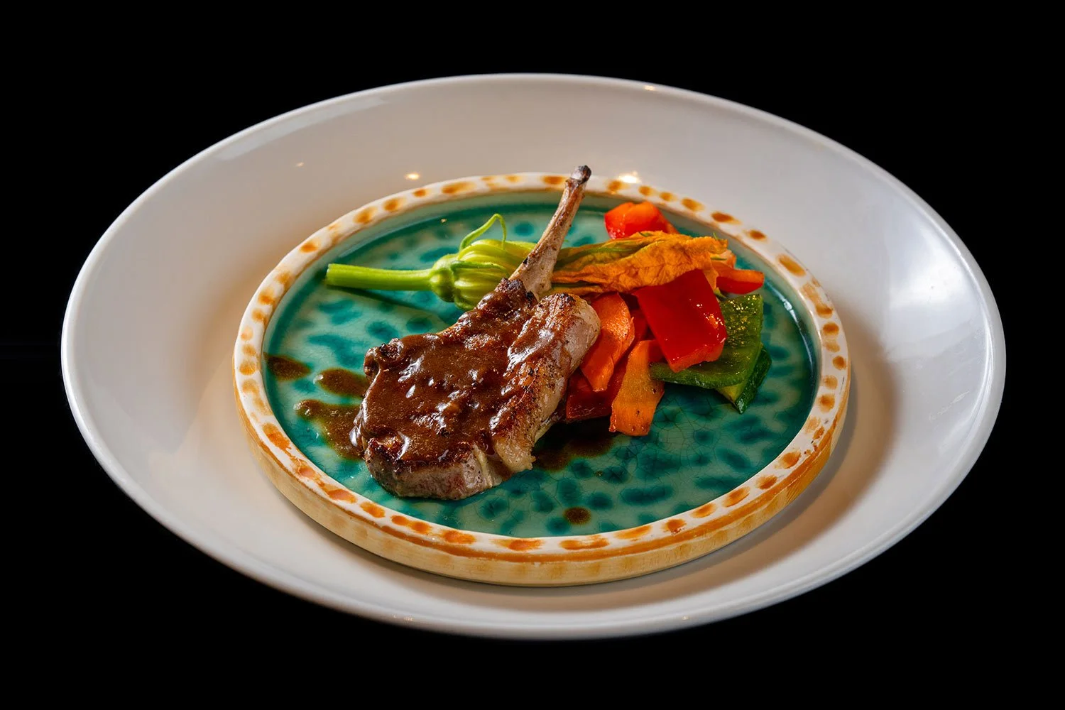 Kleines Stück Fleisch mit Sauce auf einem Teller, dazu bunte gedünstete Gemüsestücke, alles auf einem tiefblauen Teller auf weißem Servierteller