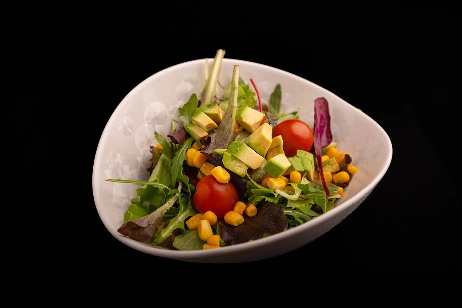 Frischer Gemüsesalat mit Cherry-Tomaten, Avocado, Süßmais, Salatblättern und Rotkohl in einer weißen Schüssel auf schwarzem Hintergrund.