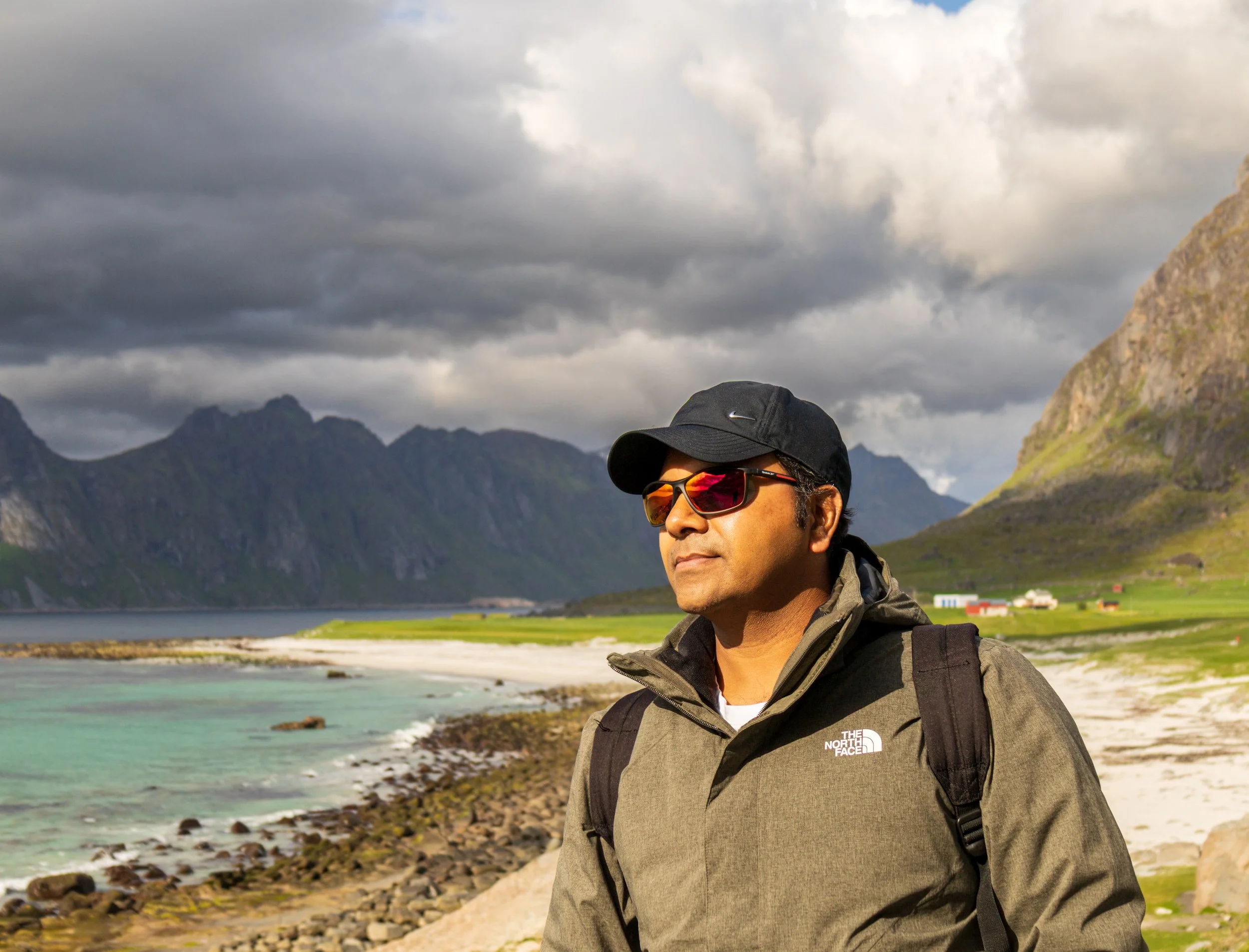 A man wearing sunglasses, a black cap, and a brown jacket with a backpack stands by a rocky shoreline with water, green fields, and mountains in the background under cloudy skies.