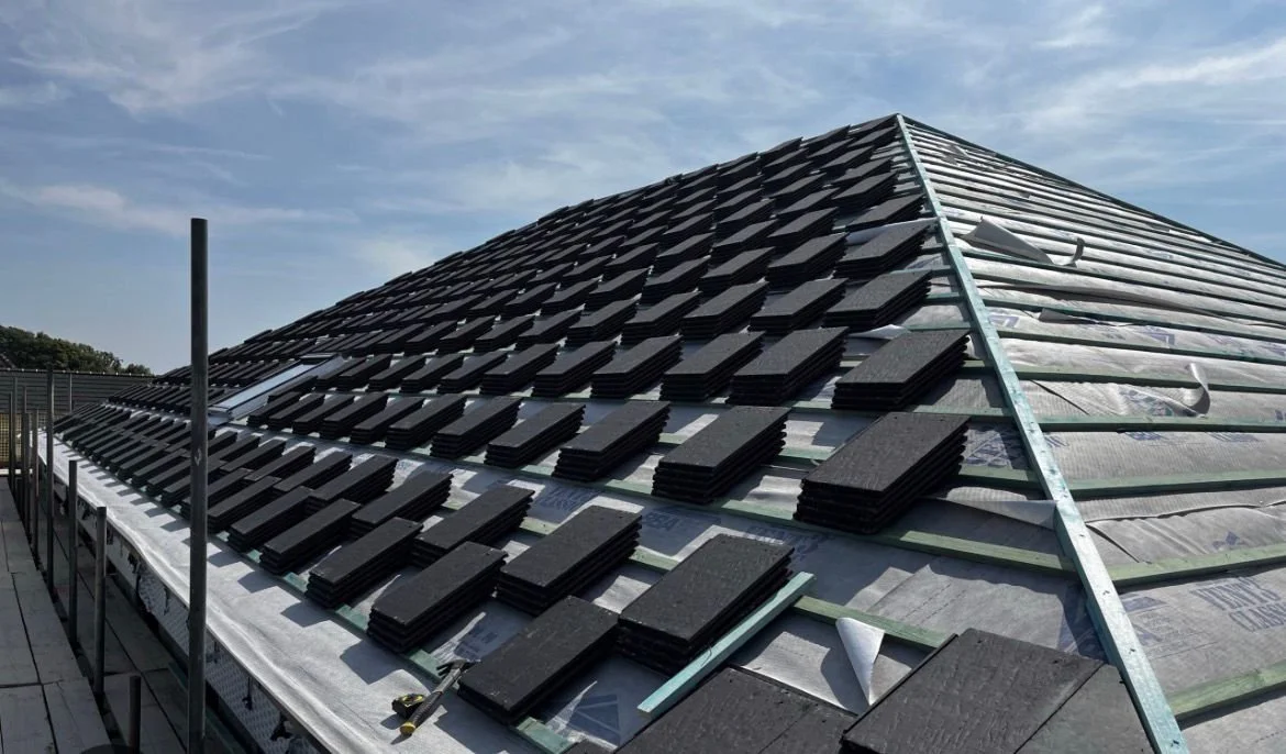 Roof under construction with black roofing tiles and metal framework, partially installed and covered with protective sheeting, against a blue sky.