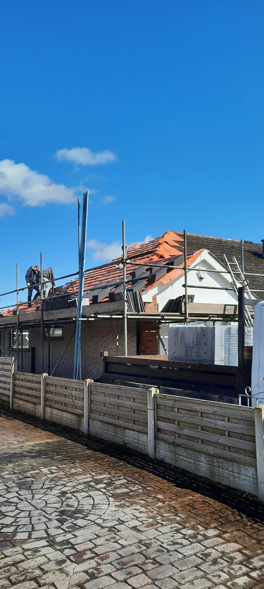 Construction workers on scaffolding fixing a house roof with red tiles under a clear blue sky.