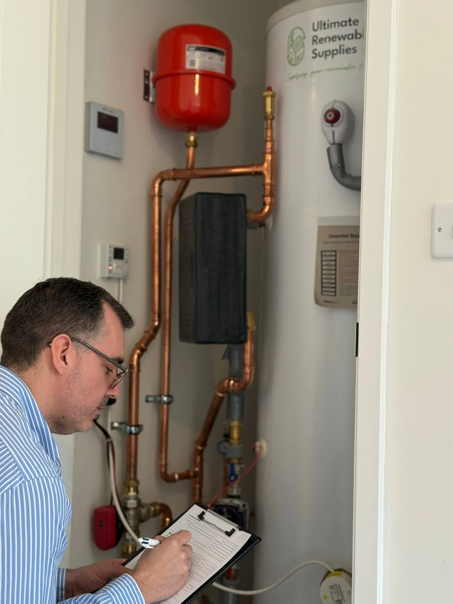 A man in a blue striped shirt writing on a clipboard while inspecting a plumbing setup with copper pipes and a water heater in a utility closet.