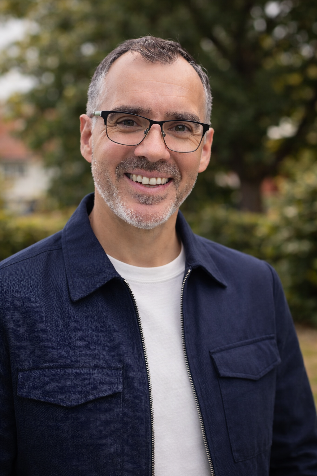 A middle-aged man with gray hair, glasses, and a beard smiling outdoors, wearing a navy jacket over a white shirt, with blurred trees in the background.