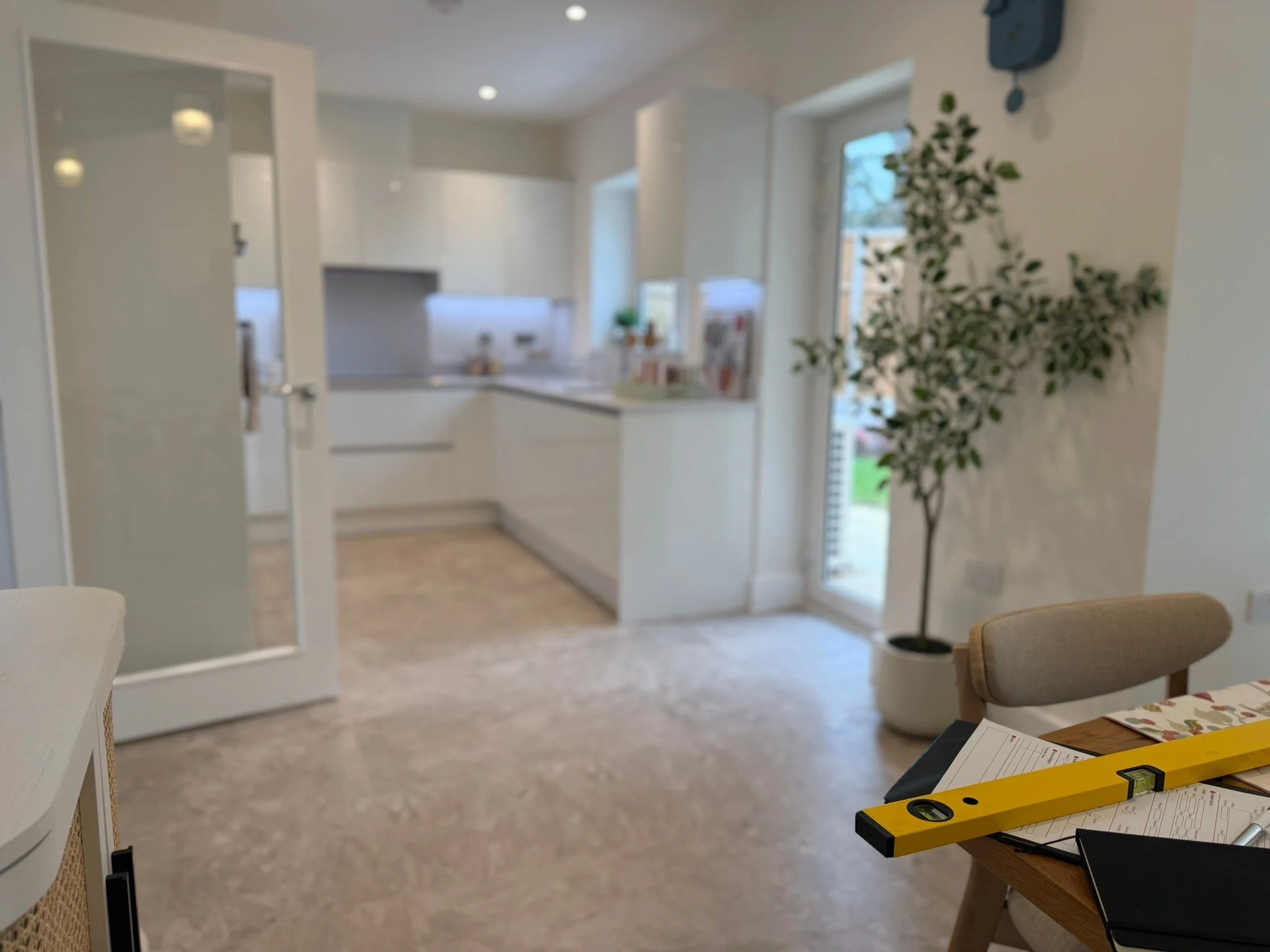 A view of a bright, modern kitchen with white cabinets, a white countertop, and multiple windows with a view outside. There is a potted plant by the window and a level on a wooden table in the foreground. The image is slightly out of focus.