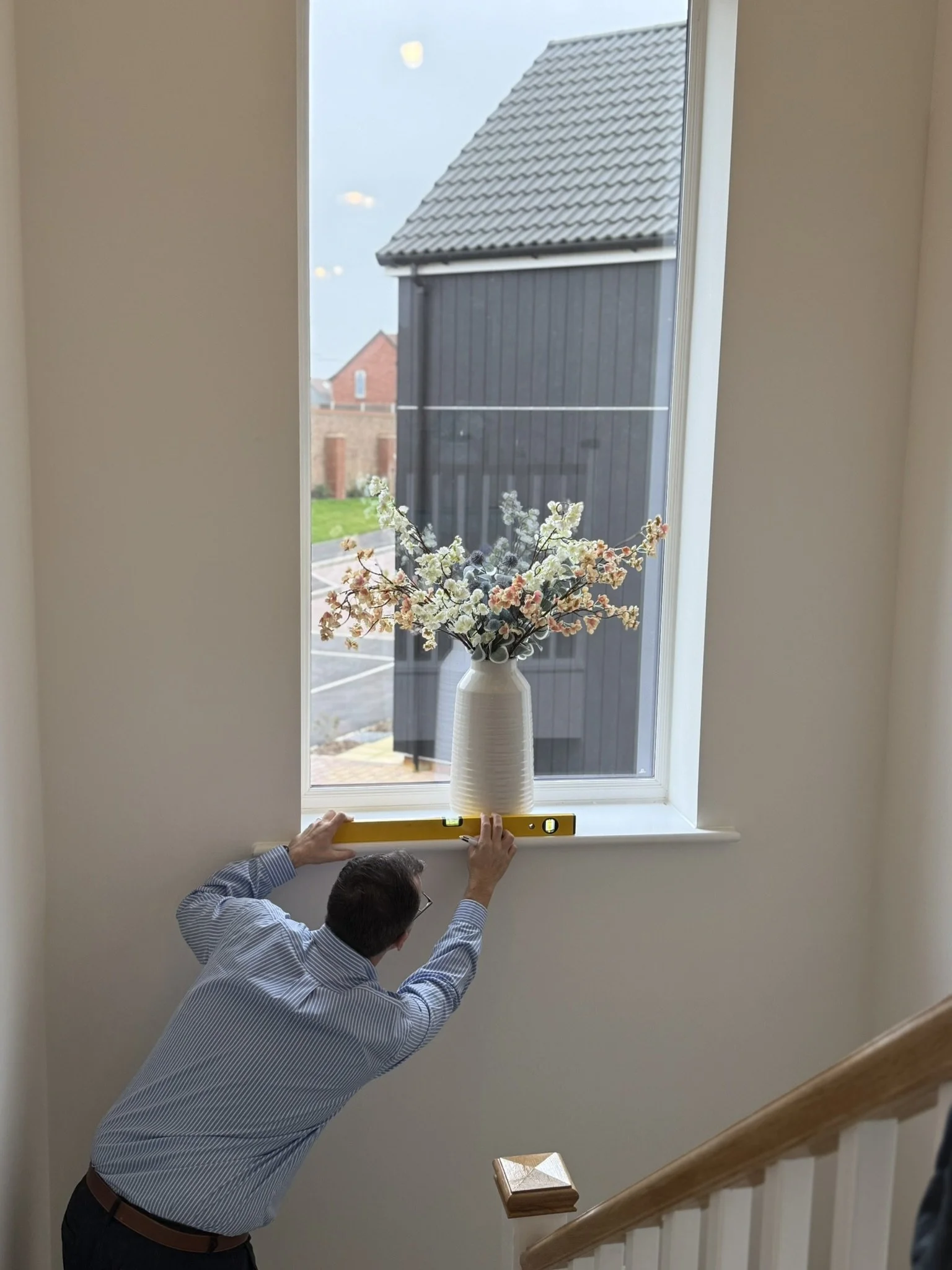 A man is installing a white vase with artificial flowers on a window sill, while using a level tool to ensure proper alignment.