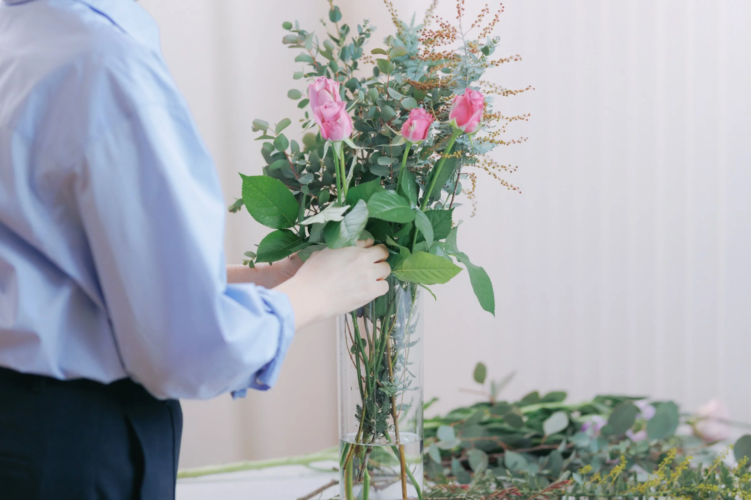 ピンクのバラの花束を持つ人のそばにいる花瓶に飾られた緑色の葉と花を含む花束の写真