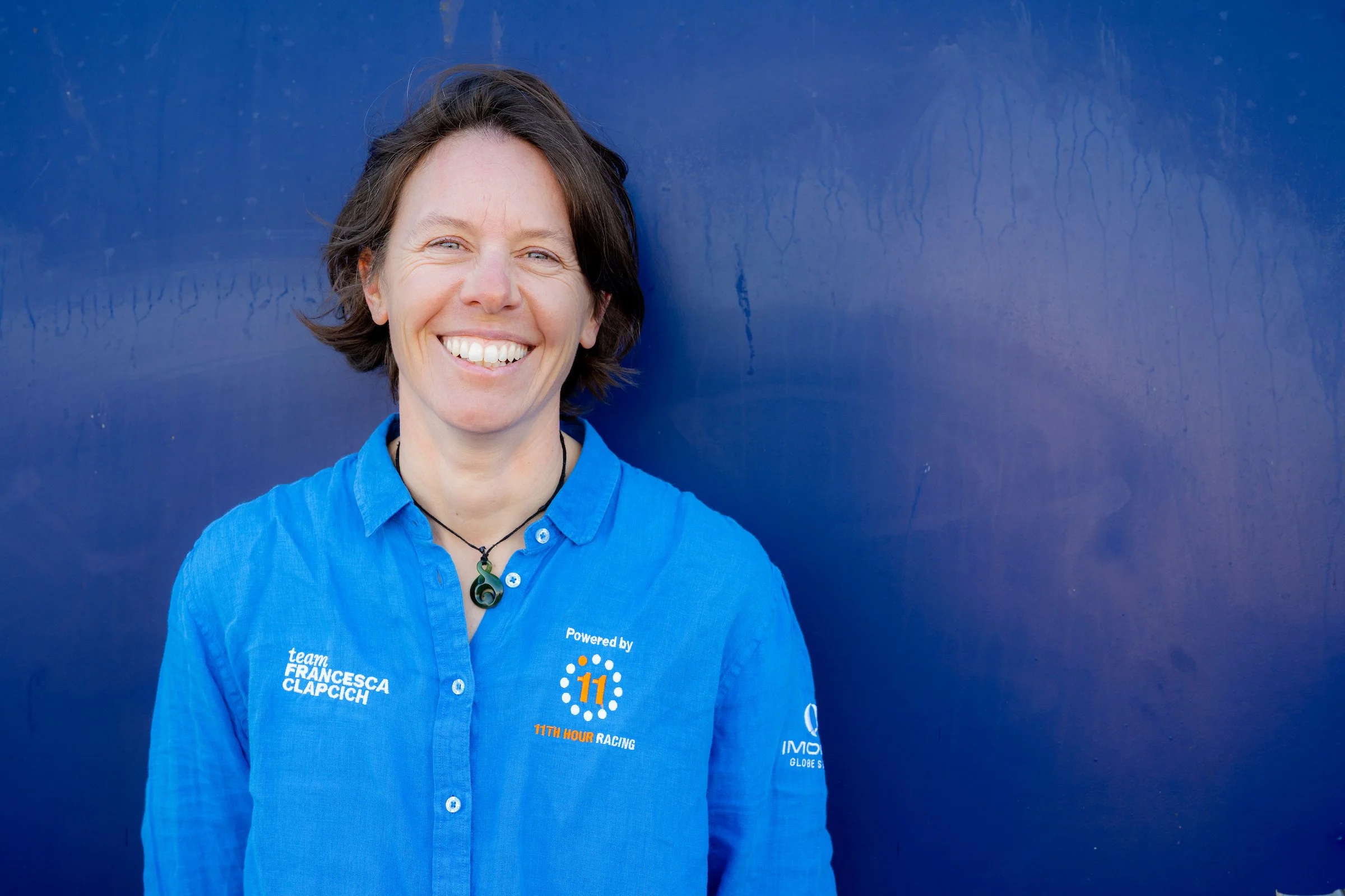 Una donna dai capelli castani corti che sorride, con indosso una camicia blu abbottonata con dei loghi, in piedi su uno sfondo blu.