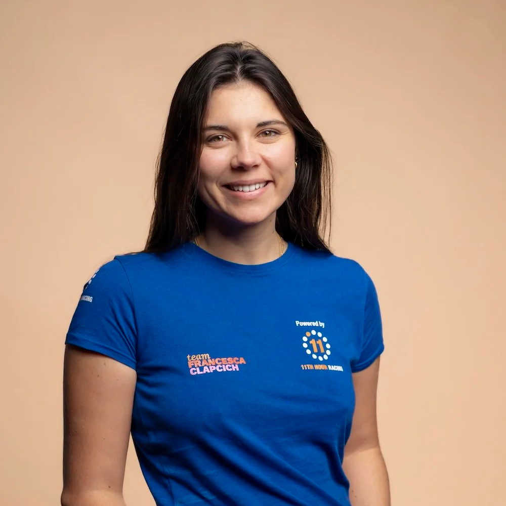 Smiling woman wearing a blue t-shirt with logos and text, standing against a peach-colored background.