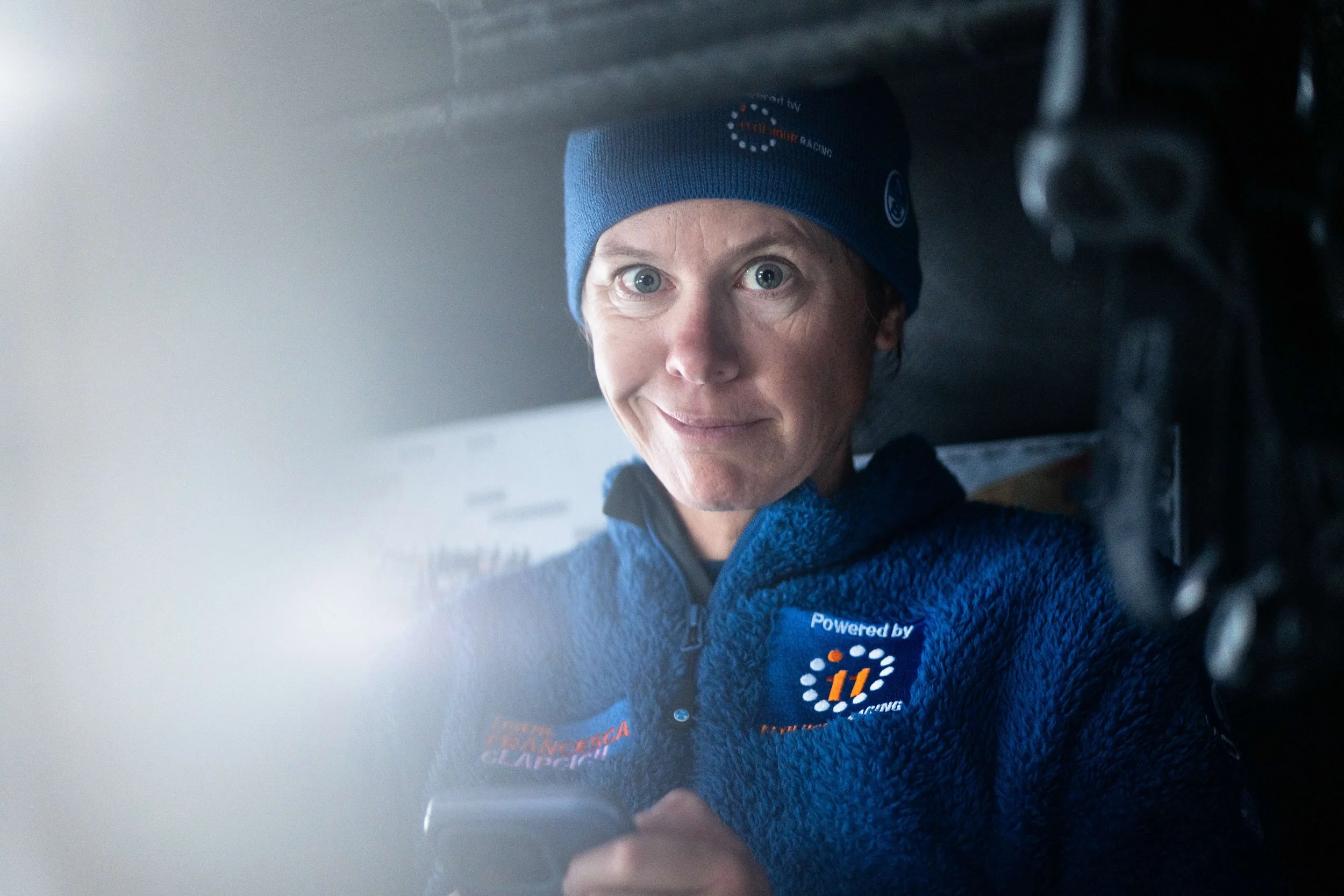 A woman wearing a blue beanie and a blue fleece jacket with various logos, looking at the camera with a slight smile.