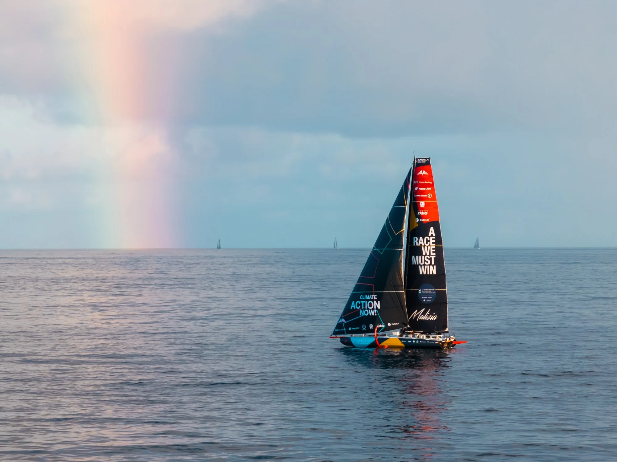 A sailboat with a black sail and colorful text and logos on it sailing on calm water, with a rainbow in the cloudy sky and other sailboats visible in the distance.