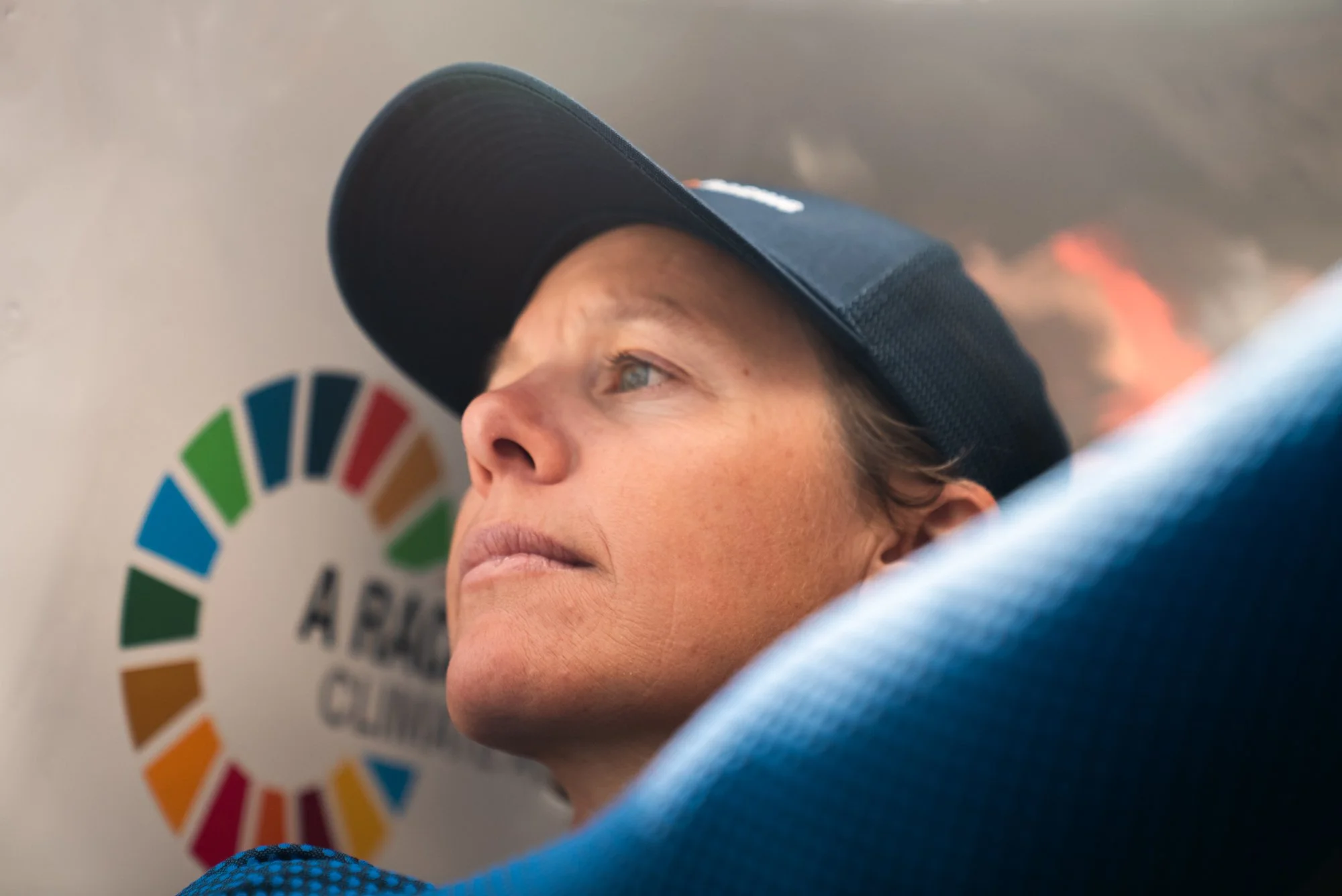 Close-up of a woman with light skin, wearing a black cap, resting her head against a blue surface, with a United Nations Sustainable Development Goals logo in the background.