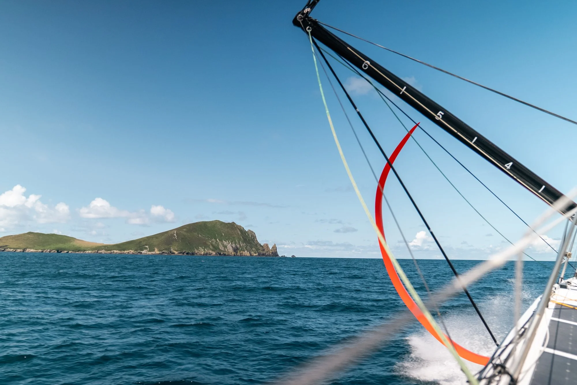 Sailing boat racing in open ocean near a green island with hills, under a clear blue sky.