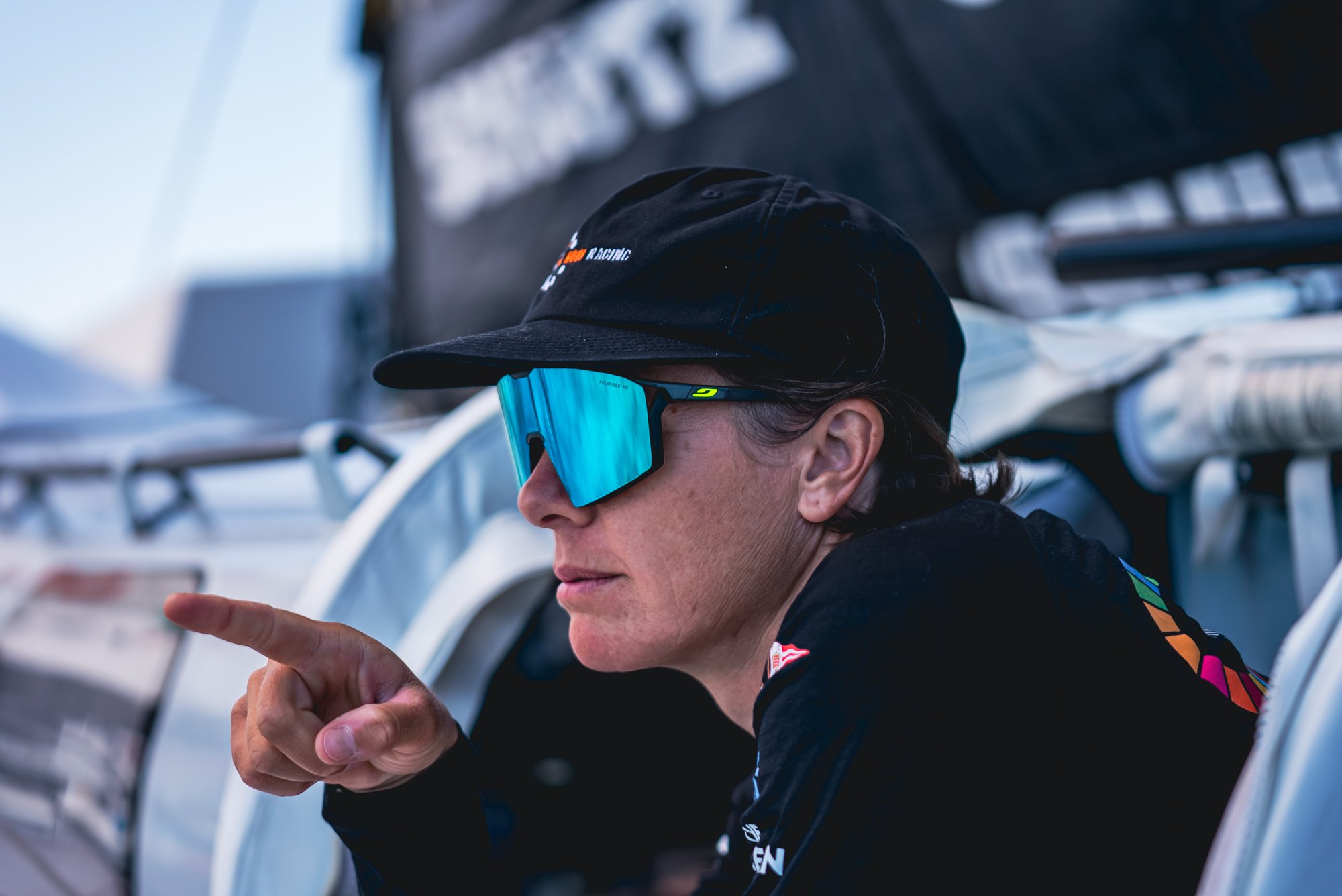 A woman wearing sunglasses, a black cap, and a black sports jacket, sitting on a race car. She is pointing to her left.