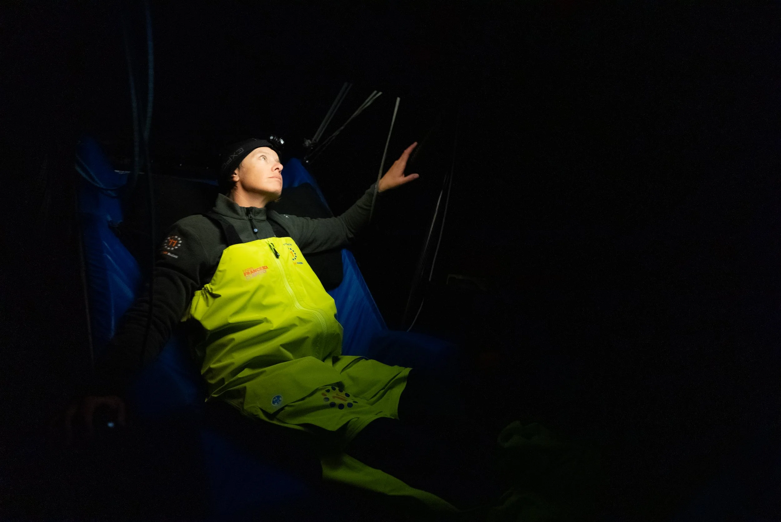 A person wearing a yellow and black jacket, sitting in a dark environment with a headlamp, reaching out with one hand.