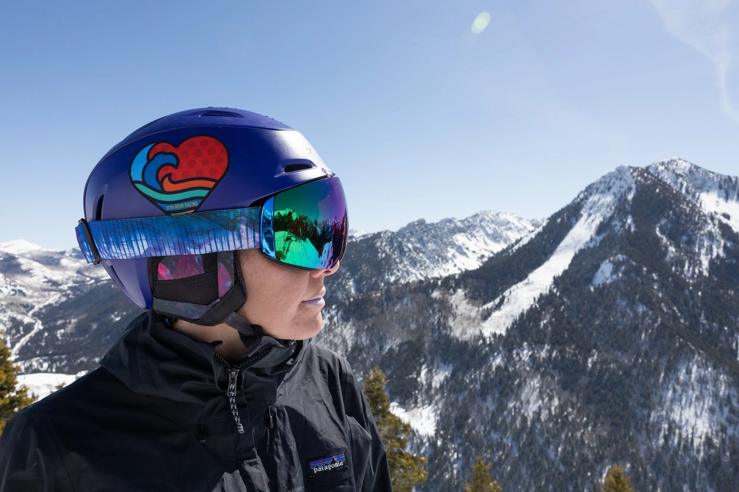 A person wearing a blue helmet with a colorful logo and reflective ski goggles, standing in snowy mountain terrain under a clear blue sky.