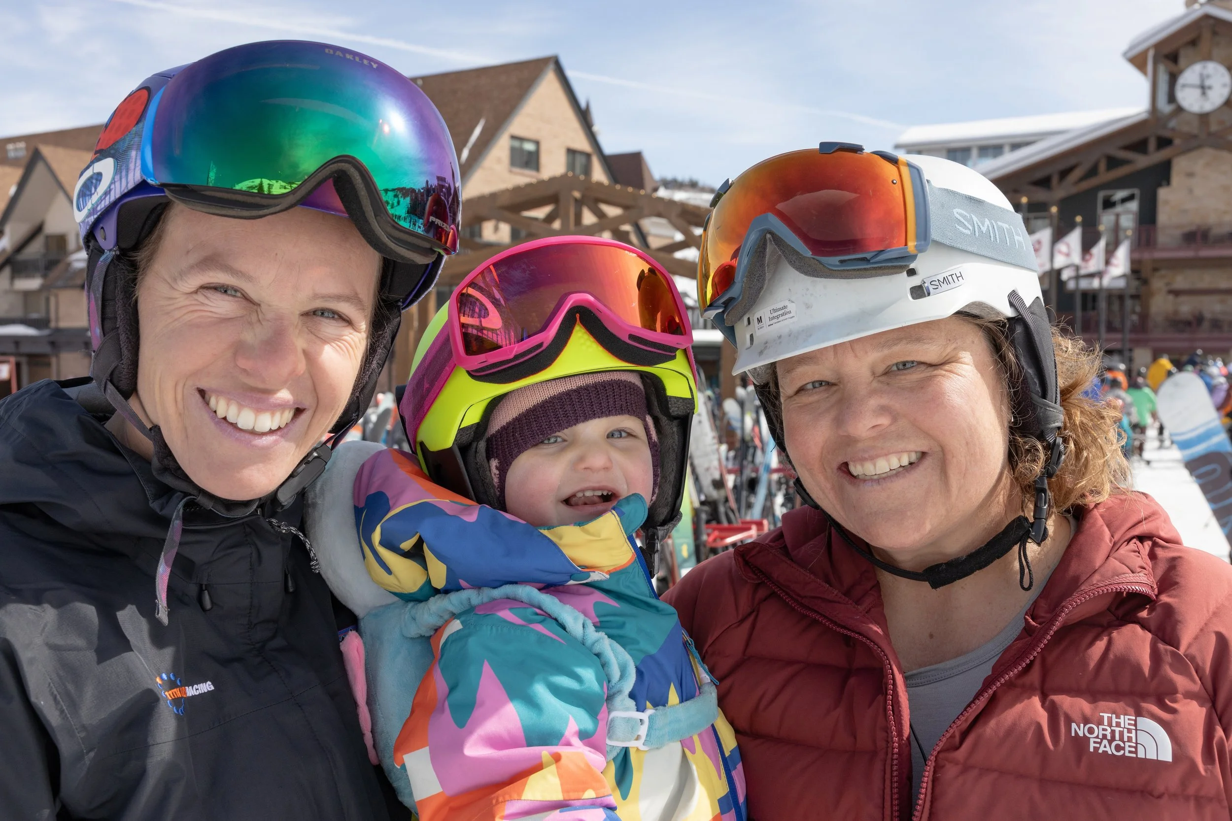 Three people in ski gear, two women and a young girl, smiling at a ski resort with snow and ski equipment in the background.