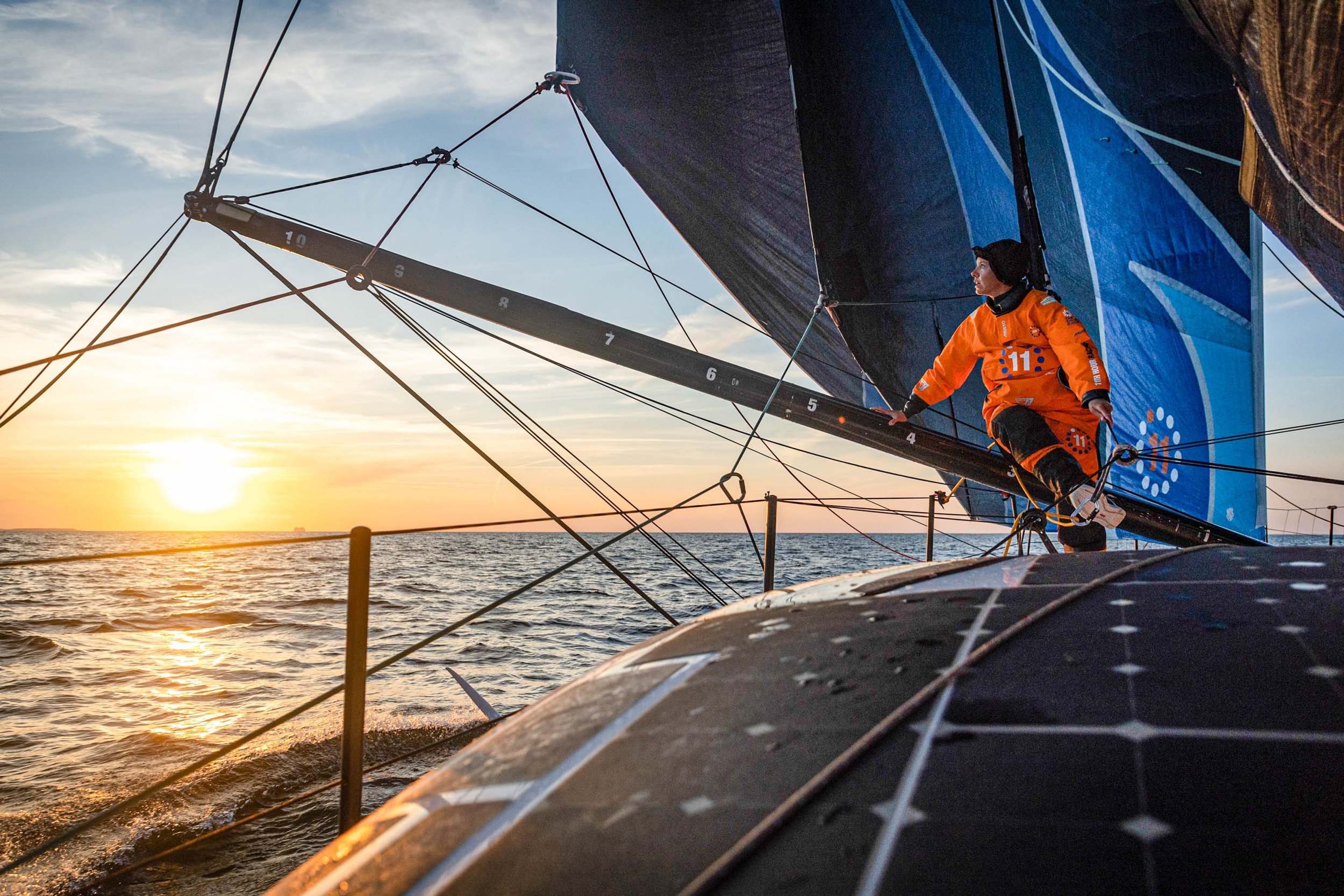 A sailor on a sailboat during sunset, wearing an orange jacket and black beanie, adjusting the rigging with the ocean and a setting sun in the background.