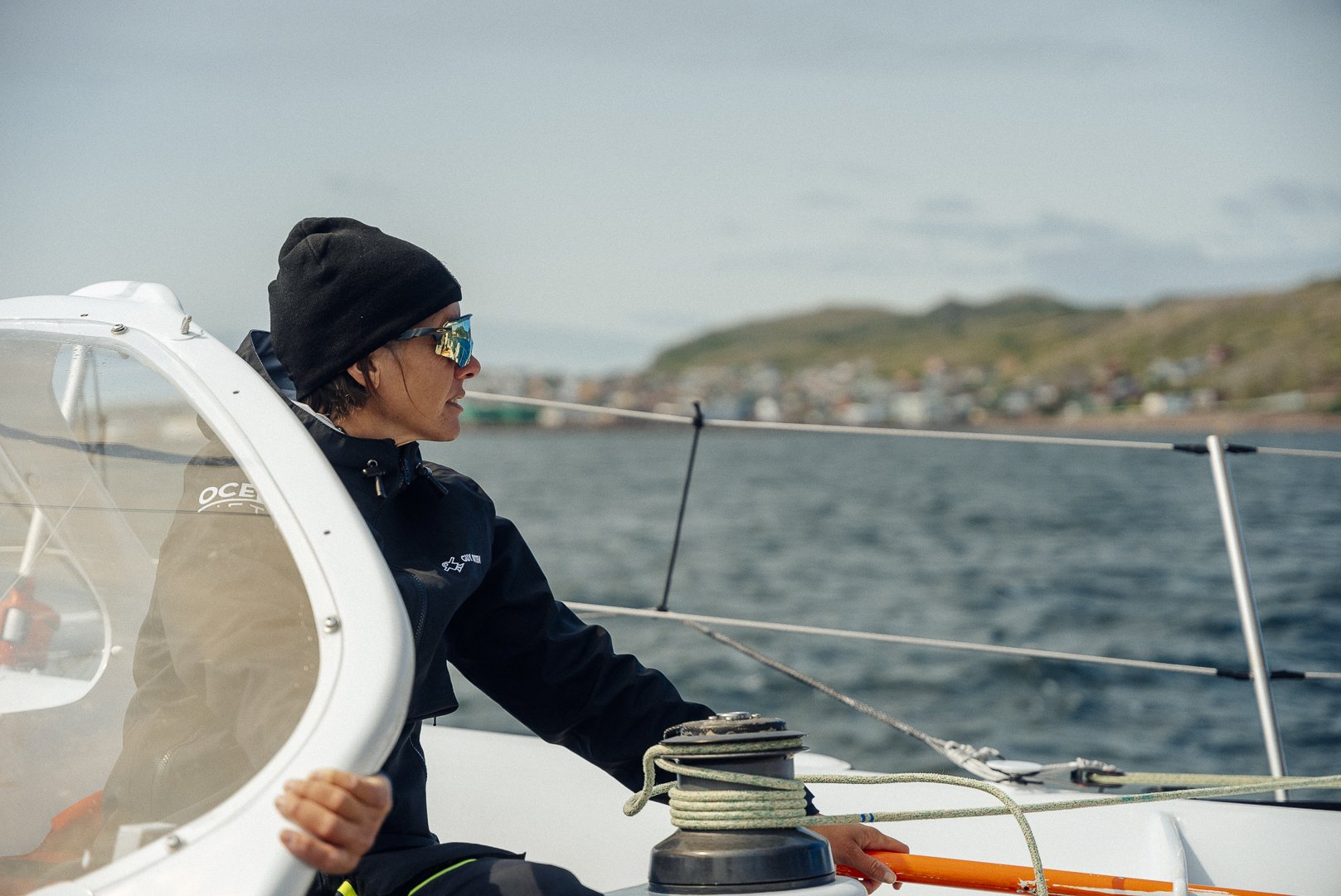 Person sailing a boat, wearing a black beanie, sunglasses, and a black jacket, on a cloudy day with water and a distant shoreline in the background.