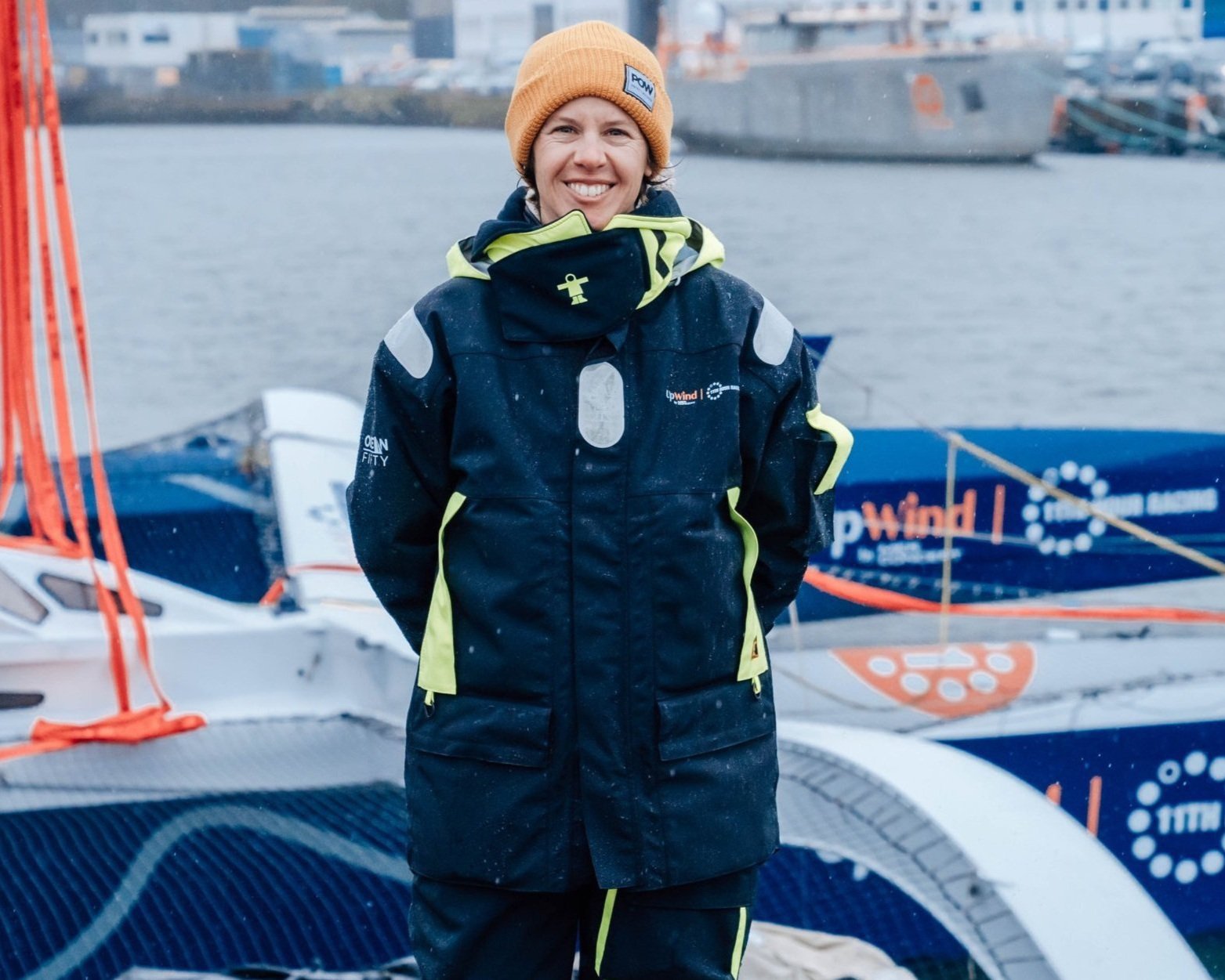 Woman in waterproof sailing gear smiling, standing near sailboats on a body of water with industrial ships in the background.