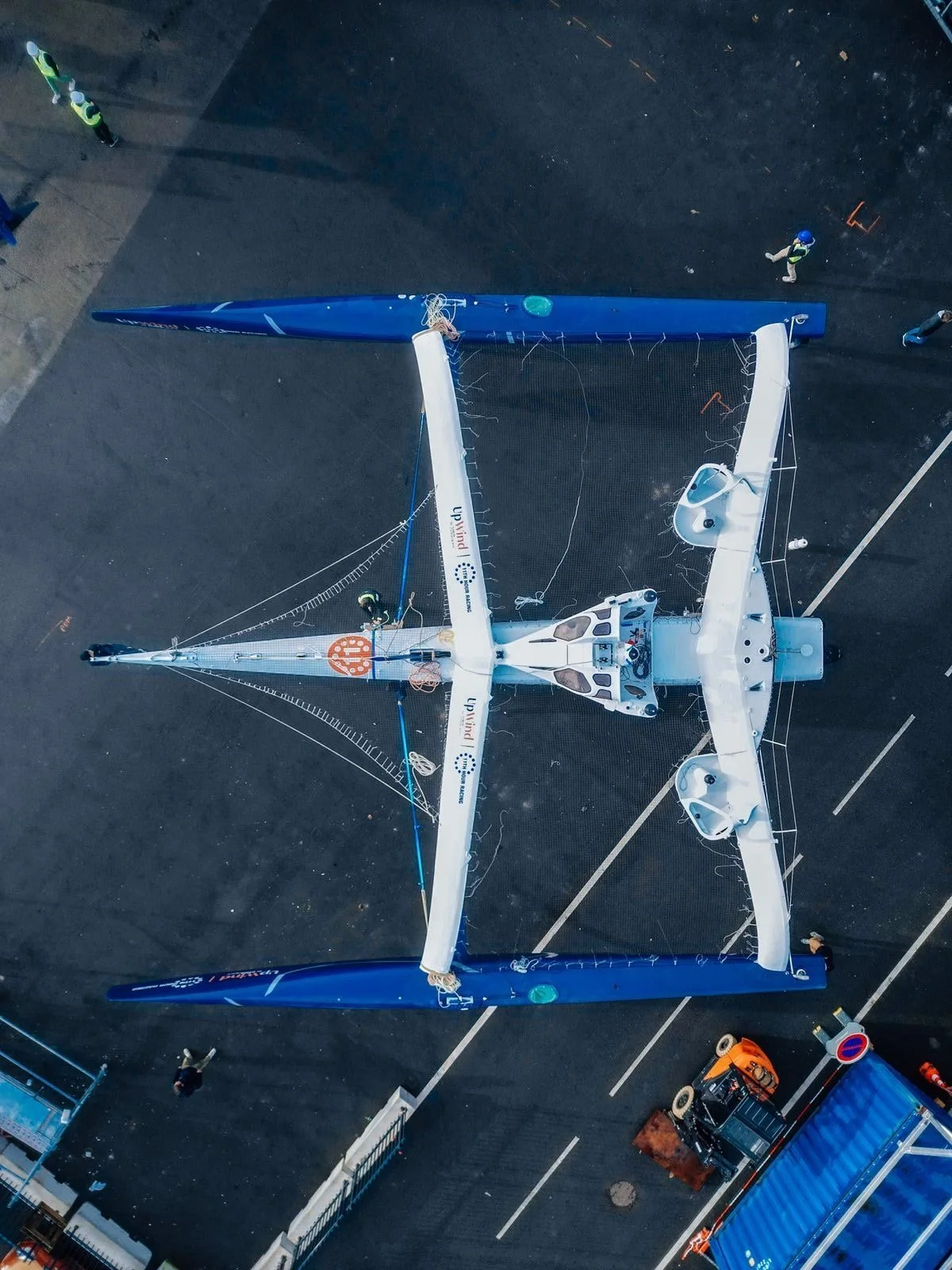 A robotic sailboat with a white and blue hull and tall sails, situated on a paved surface with several people nearby.